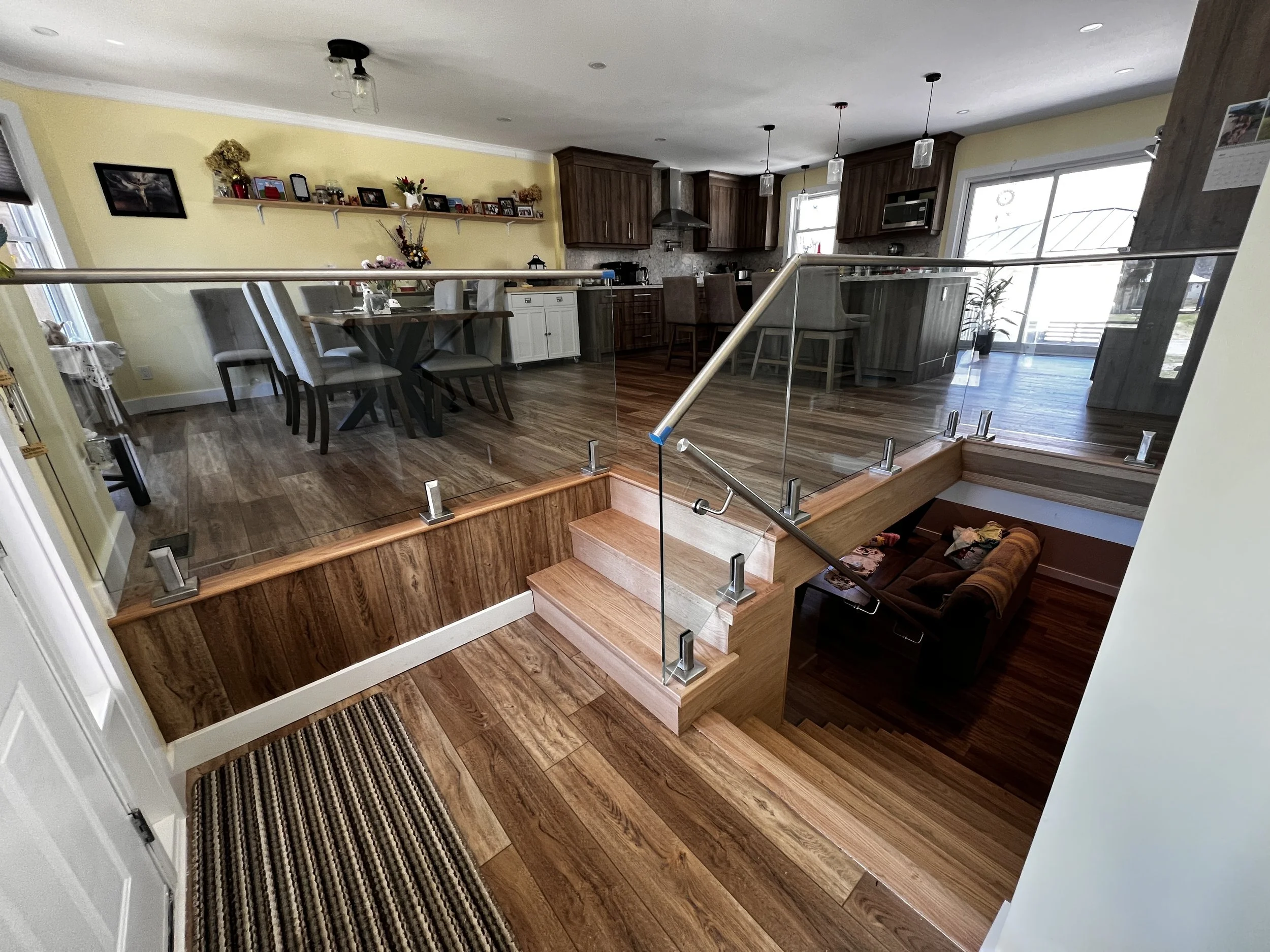 Interior view of a home with a wooden staircase topped by glass railing, leading up to a dining area and kitchen. The space includes wooden floors, a dining table with chairs, and kitchen cabinets. Large windows allow natural light to fill the room.