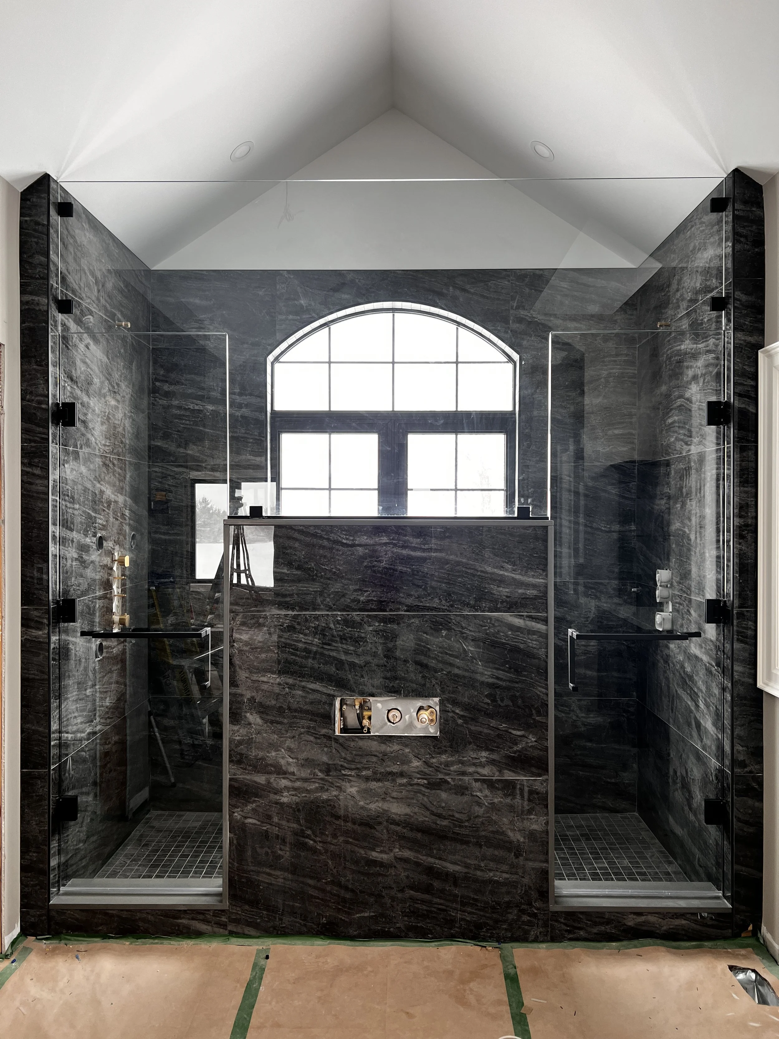 Modern shower with black marble walls and glass doors, large arched window, and black fixtures.