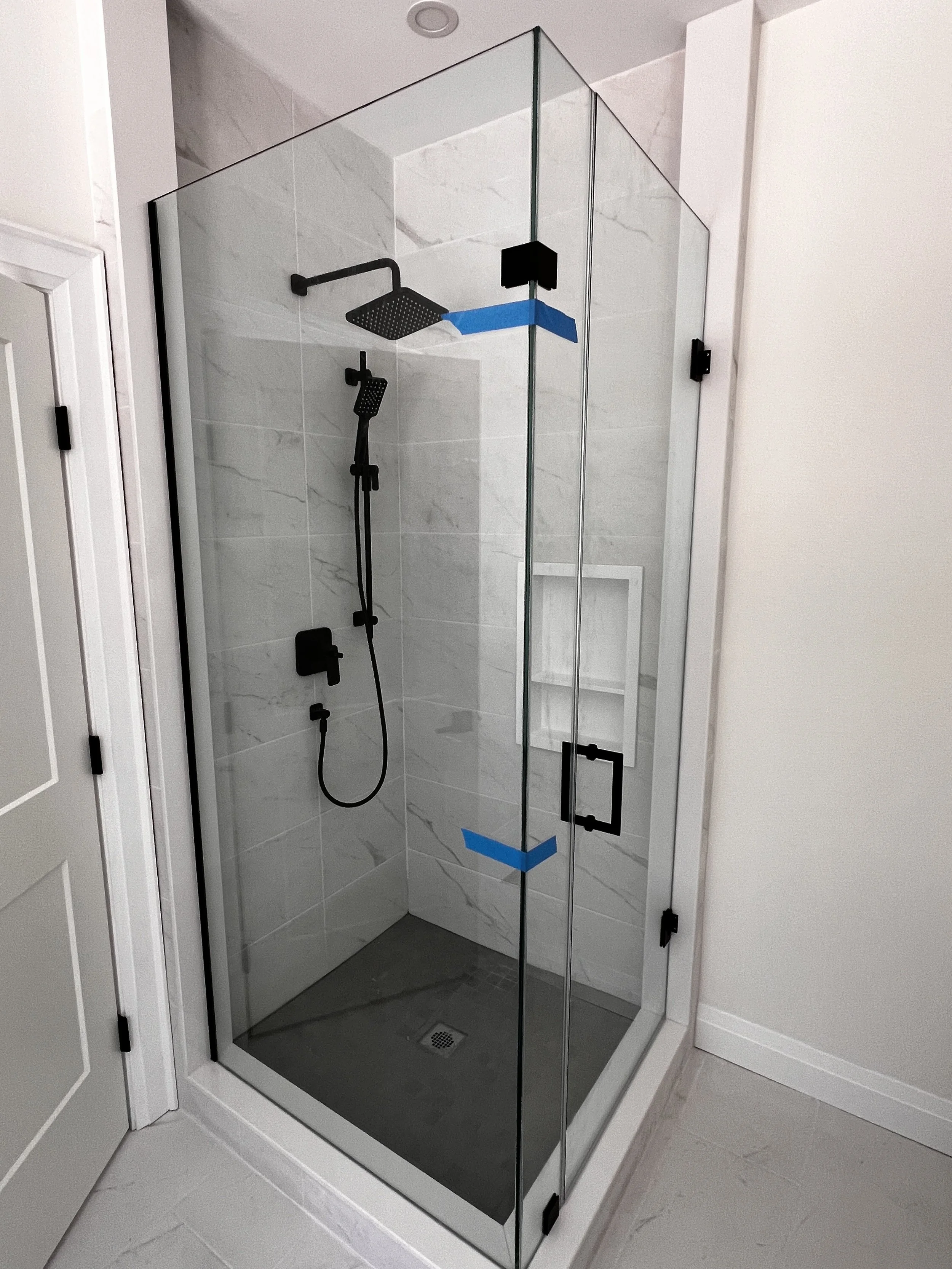 A modern glass shower enclosure with black fixtures, including a showerhead and handheld spray, installed in a bathroom with marble tiles and a small built-in shelf.