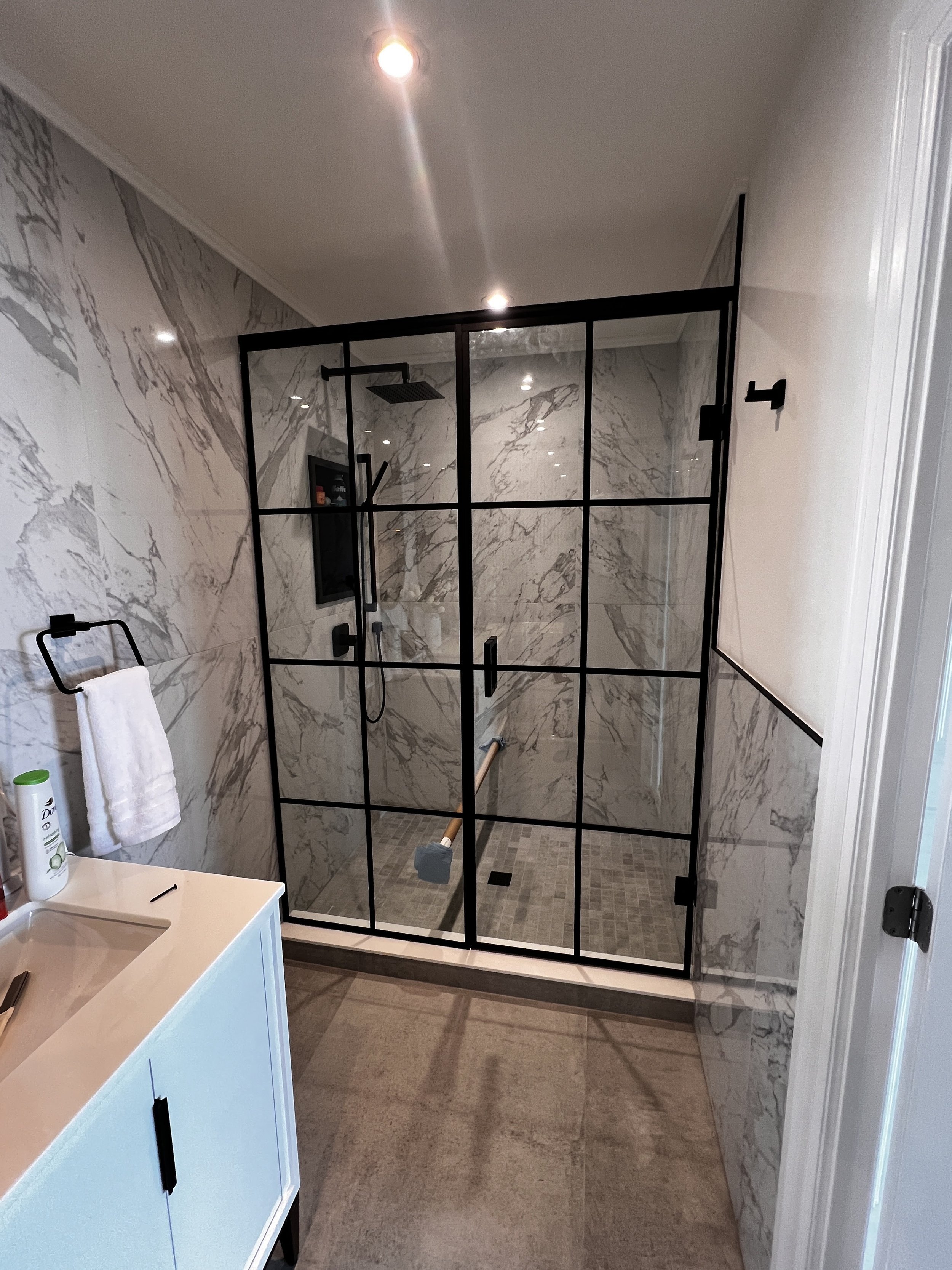 Modern bathroom with a glass-enclosed shower featuring black metal framing, marble walls, a rain showerhead, and a wooden grab bar. There is a white vanity with a black handle, a towel hanging on a black towel bar, and a small shelf inside the shower with toiletries.