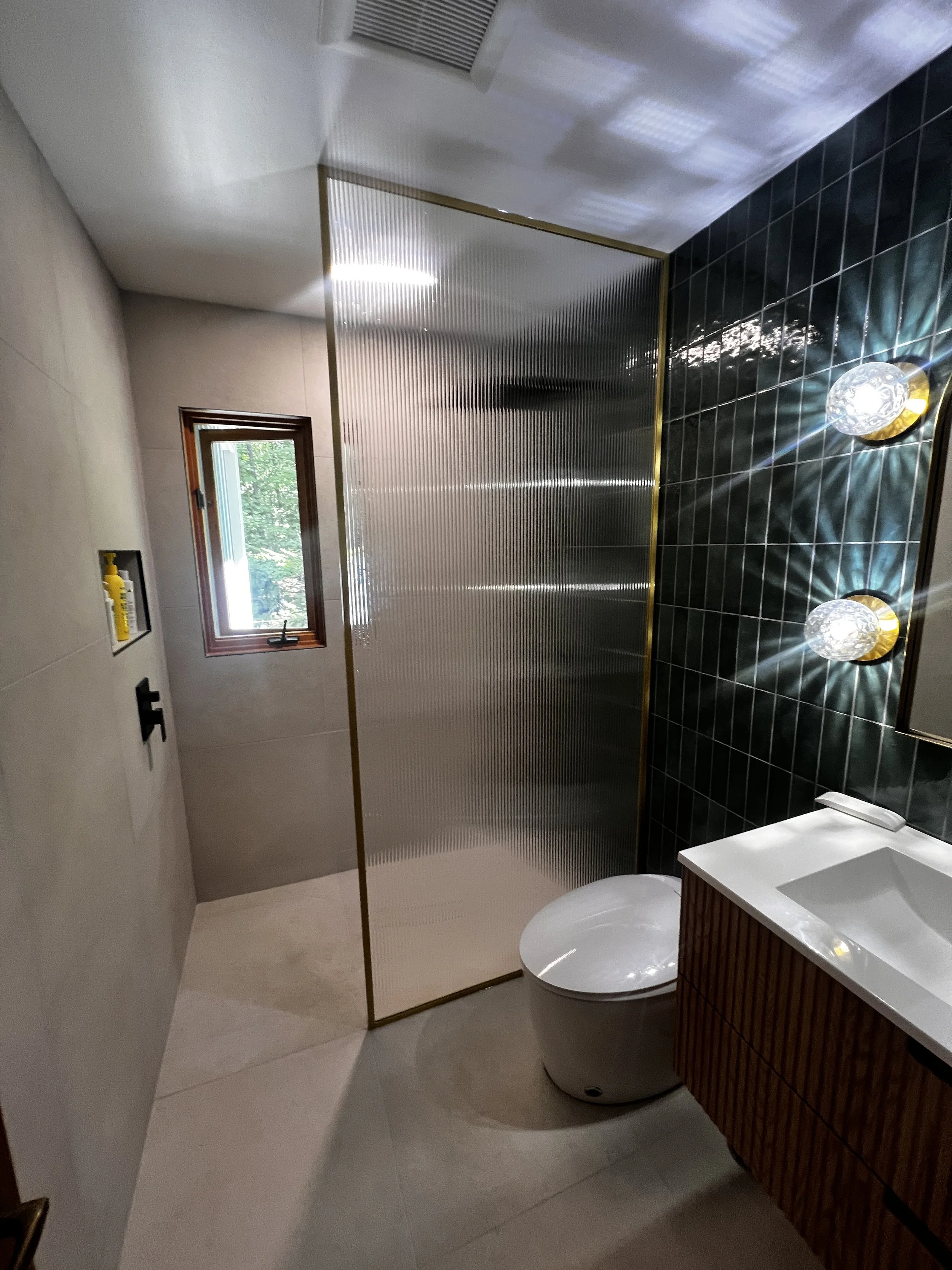 Modern bathroom with a small window, a sliding door with textured glass, a white toilet, a wooden vanity with a white sink, and black tiled walls with two round wall sconces.