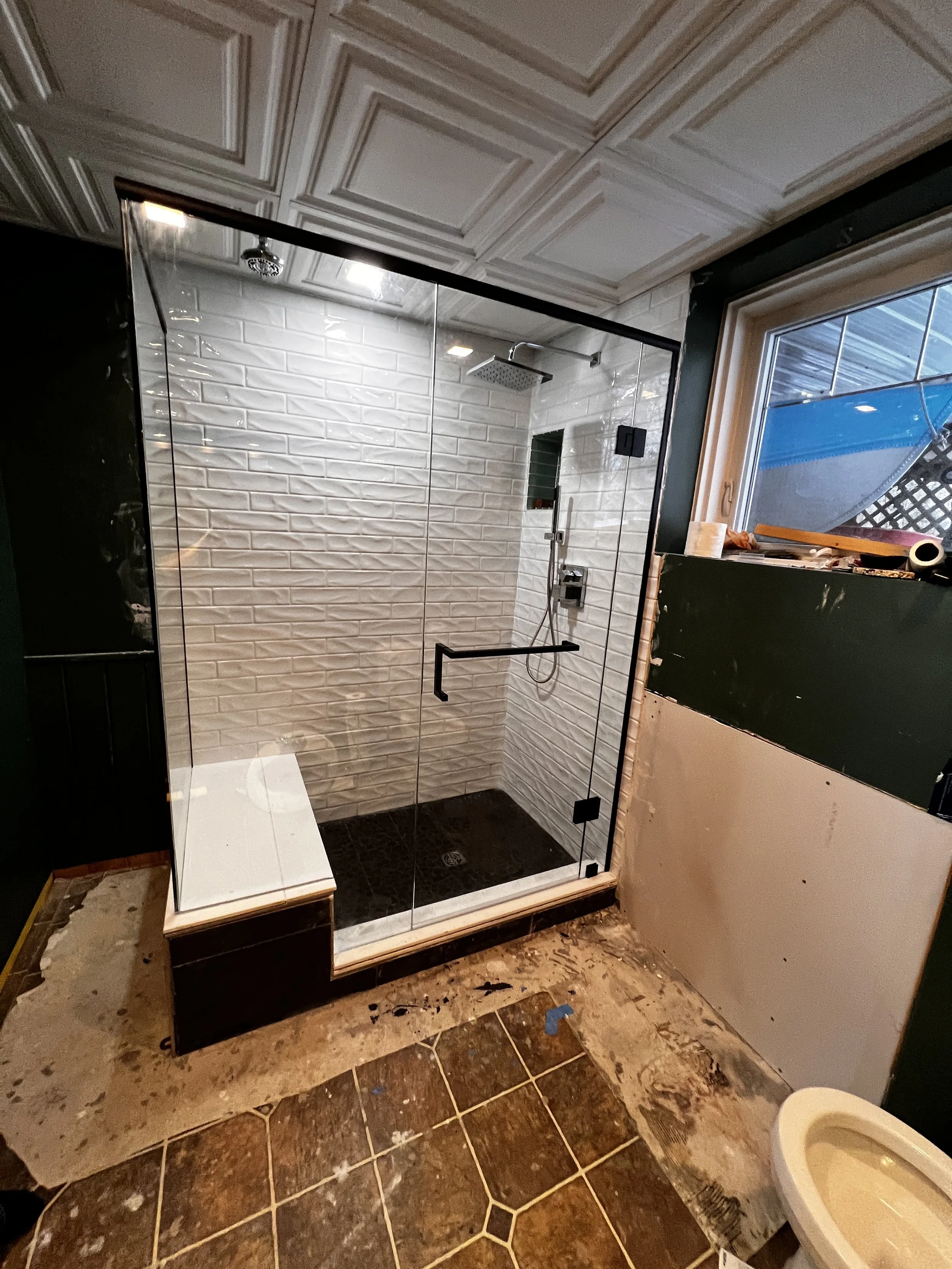 A shower enclosed with glass panels and black hardware, white subway tile walls, a rain shower head, and a black shower control panel, in a bathroom under renovation with unfinished floor and wall area.