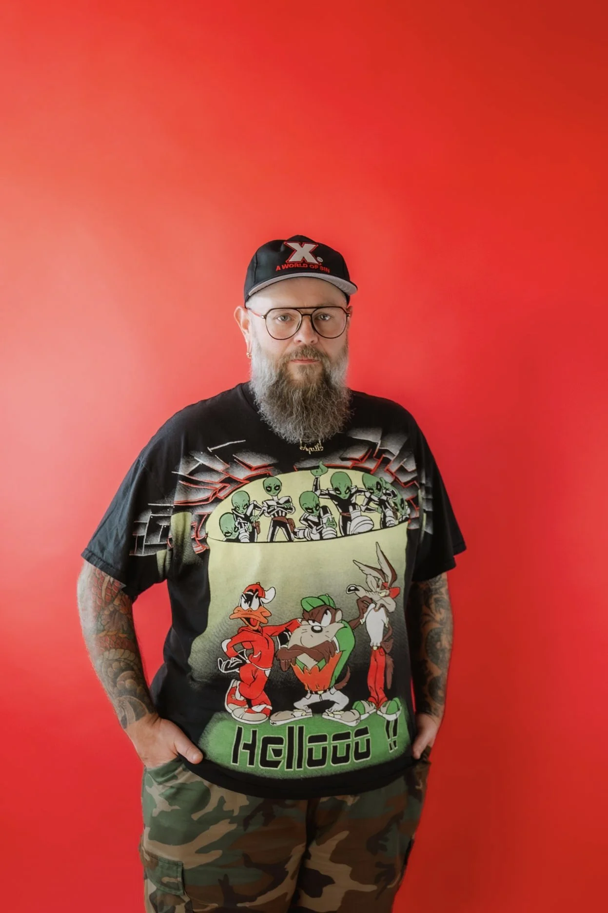 A man with glasses, a beard, and tattoos is standing against a red background. He's wearing a black T-shirt featuring cartoon characters and a black cap with the letter 'X' and the slogan 'A WORLD OF SIN.'