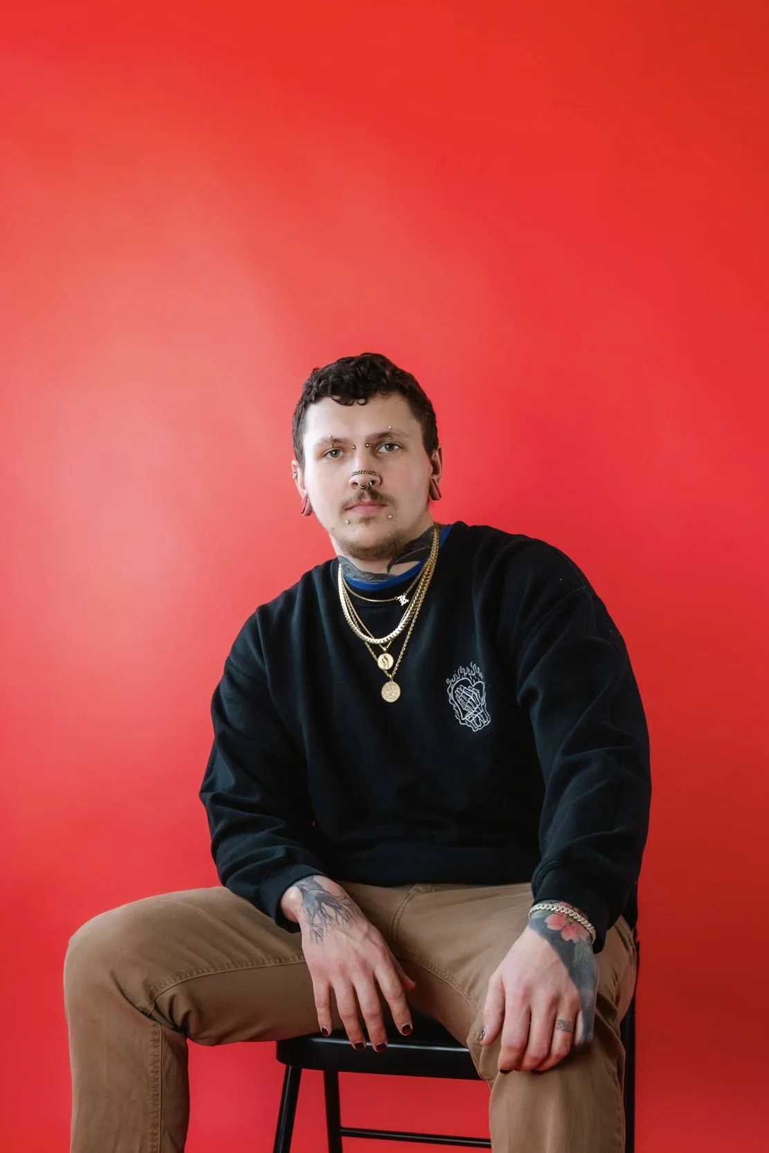 A man with tattoos, piercings, and a dark shirt posing with his hand on his chin against a red gradient background.