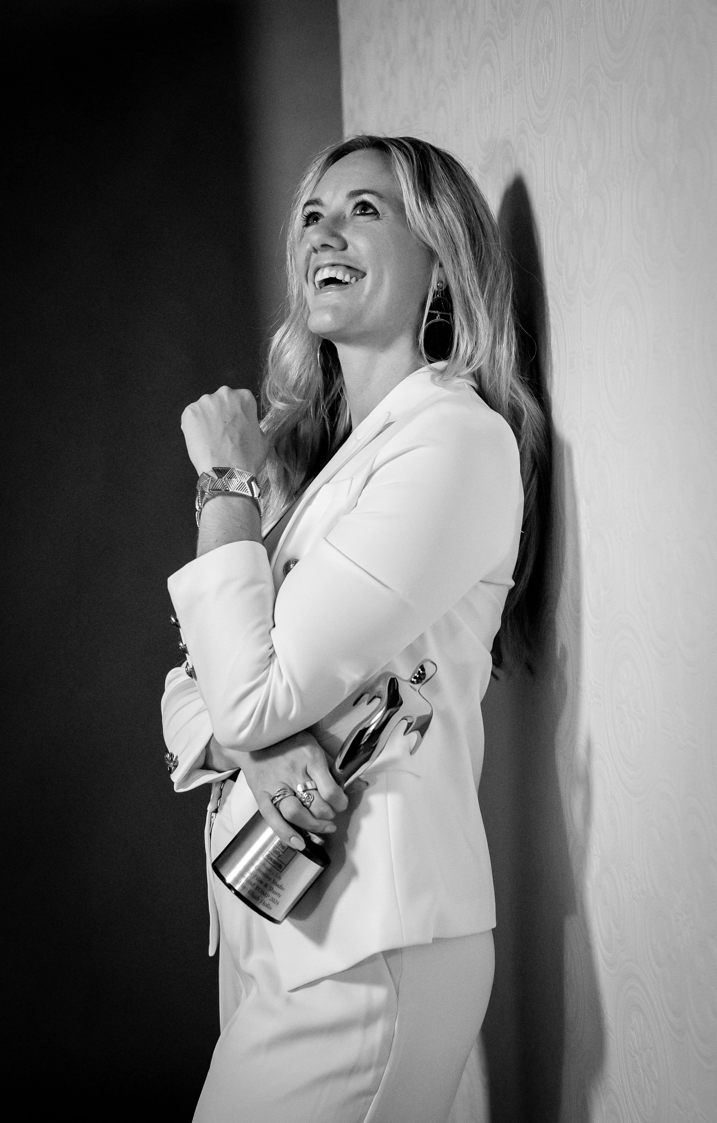 Professional shot of Ashlee Hollis sitting on a chair smiling at the camera with her legs crossed and one hand propped under her face.