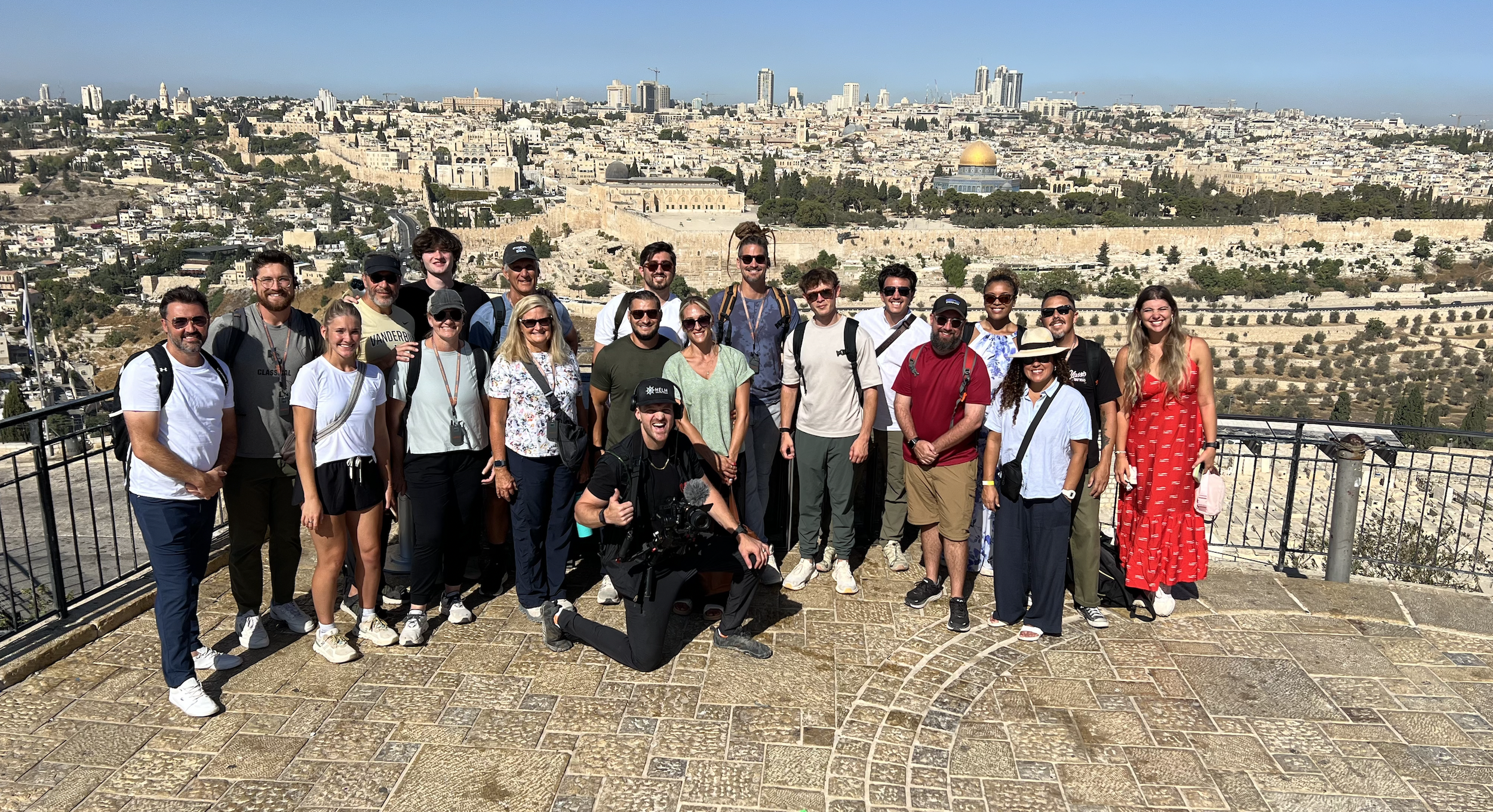 Elijah Hollis holds his camera on a balcony in Jerusalem, Israel with a group of people and the Dome of the Rock in the background distance.