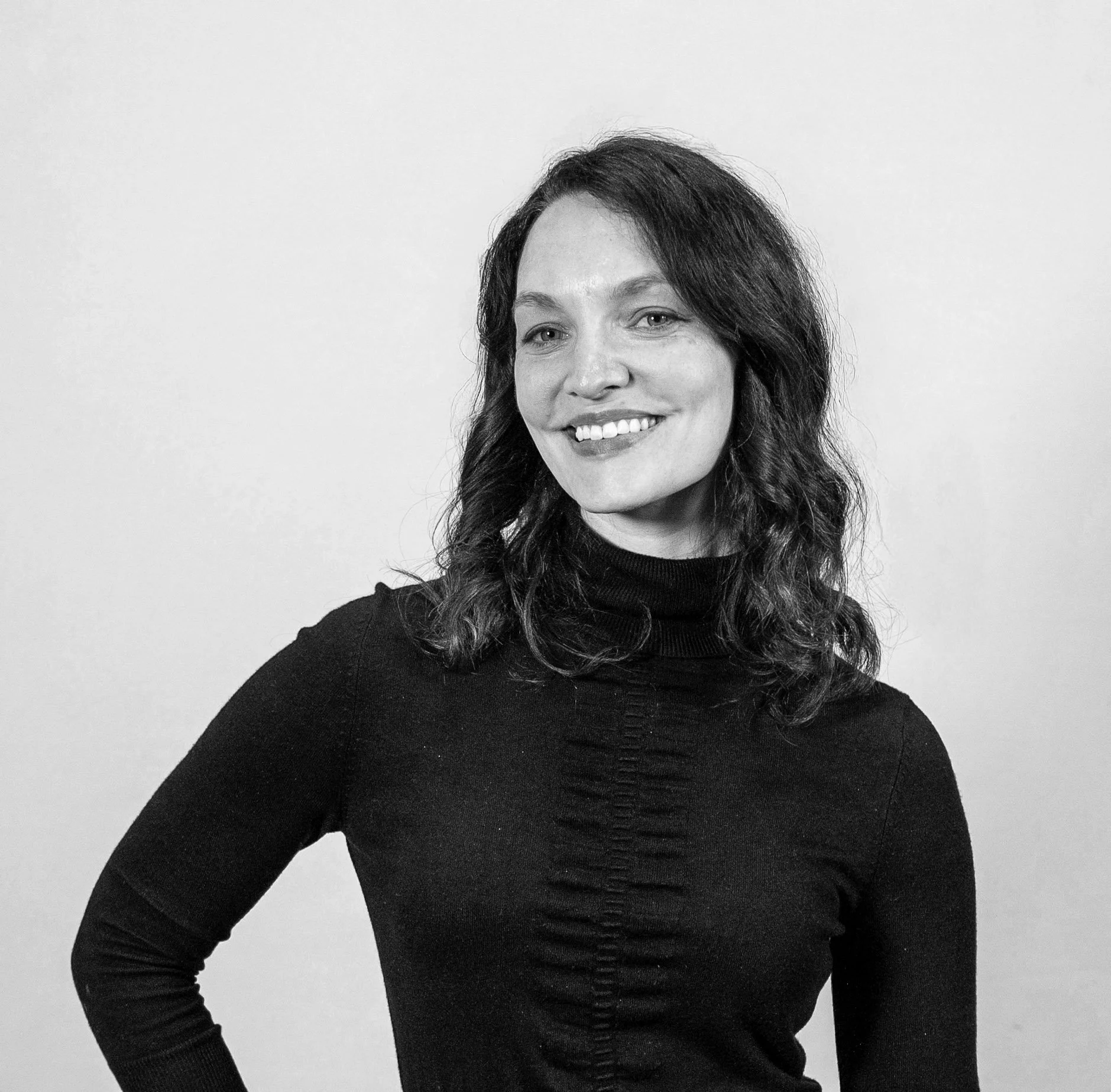 Headshot of Sara Norcott standing to the side with her head angled towards the camera as her right arm grabs her left while smiling. 