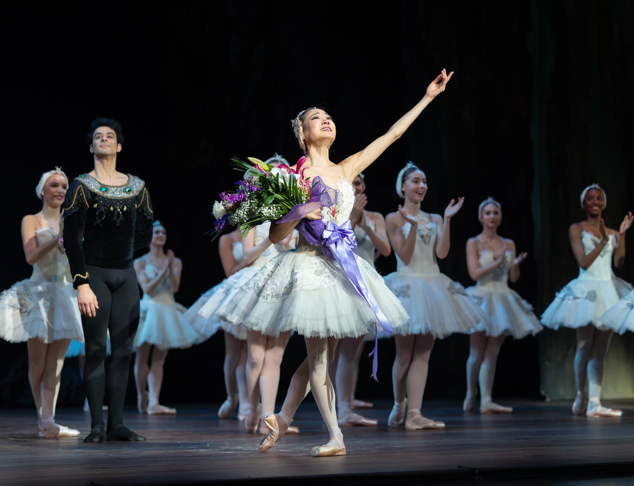 A ballerina in a Swan Lake costume stands on a stage with dancers behind her and holds flowers while the other arms is raised as she smiles.