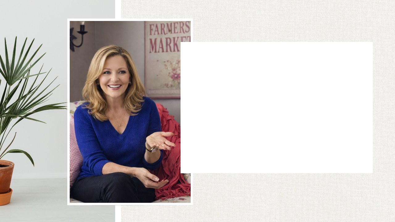 A woman with shoulder-length blonde hair, wearing a royal blue sweater, sitting on a sofa and smiling while gesturing with her right hand in a room with a sign that says 'Farmers Market' in the background.
