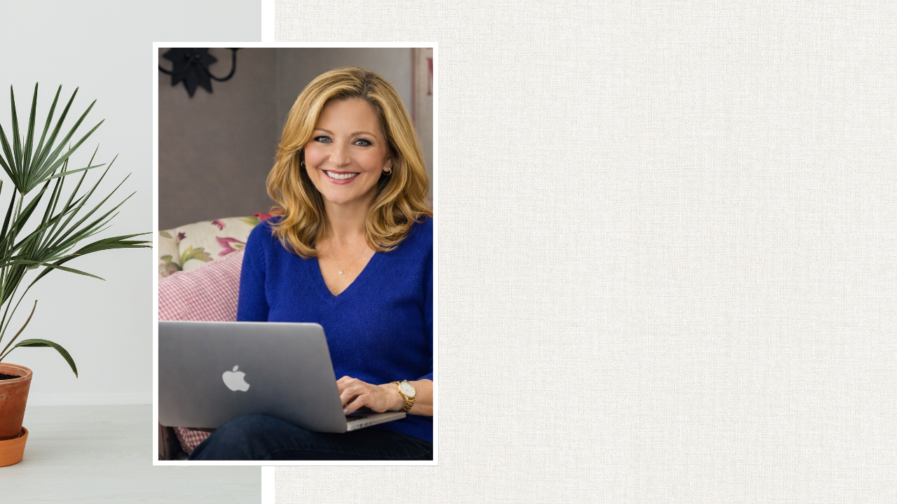 A woman with blonde hair in a blue sweater smiling while sitting with a laptop on her lap, in a cozy home setting.