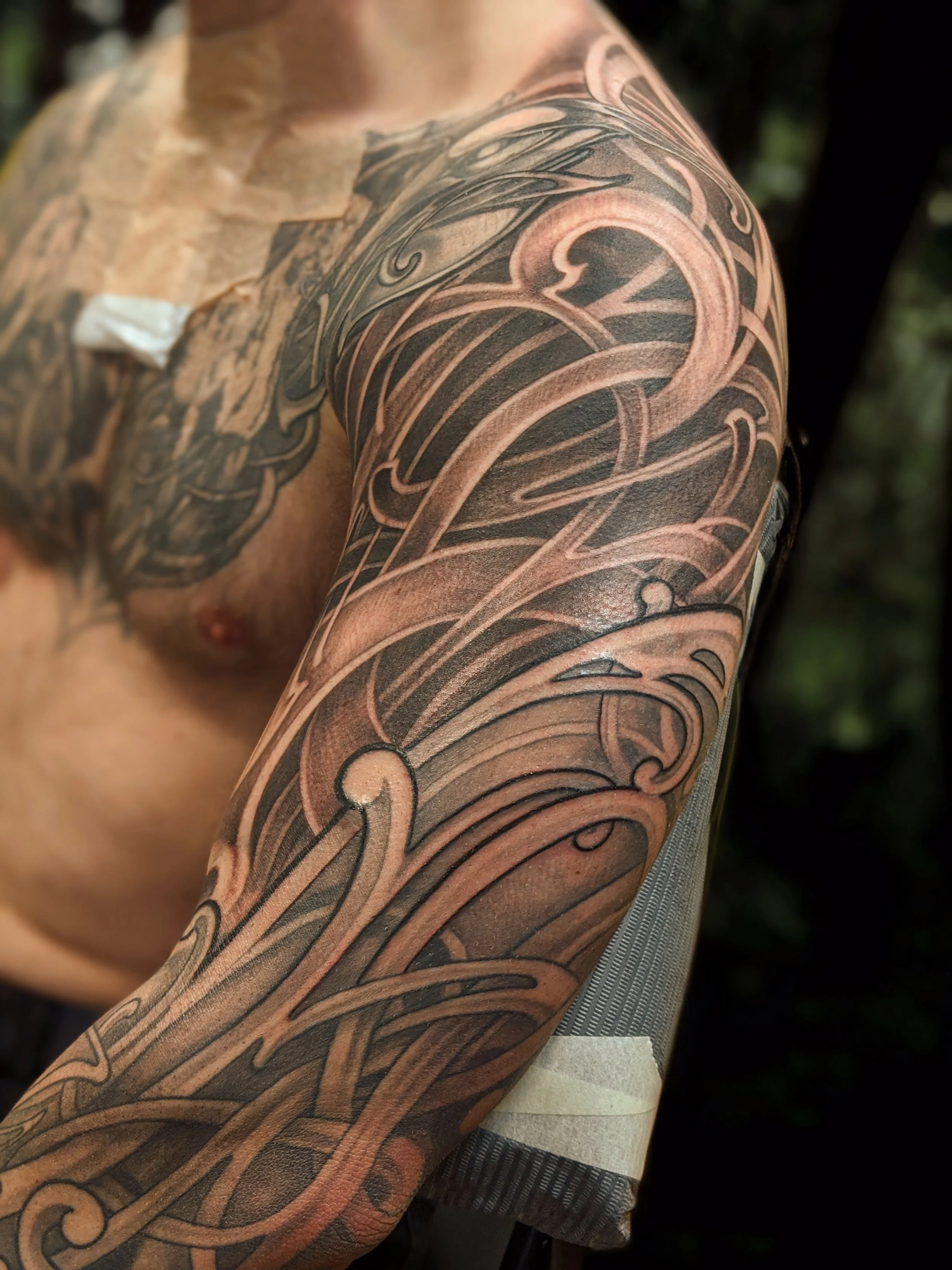 Close-up of a person's shoulder and upper arm with an intricate black and gray tattoo featuring flowing, swirling lines and abstract patterns.