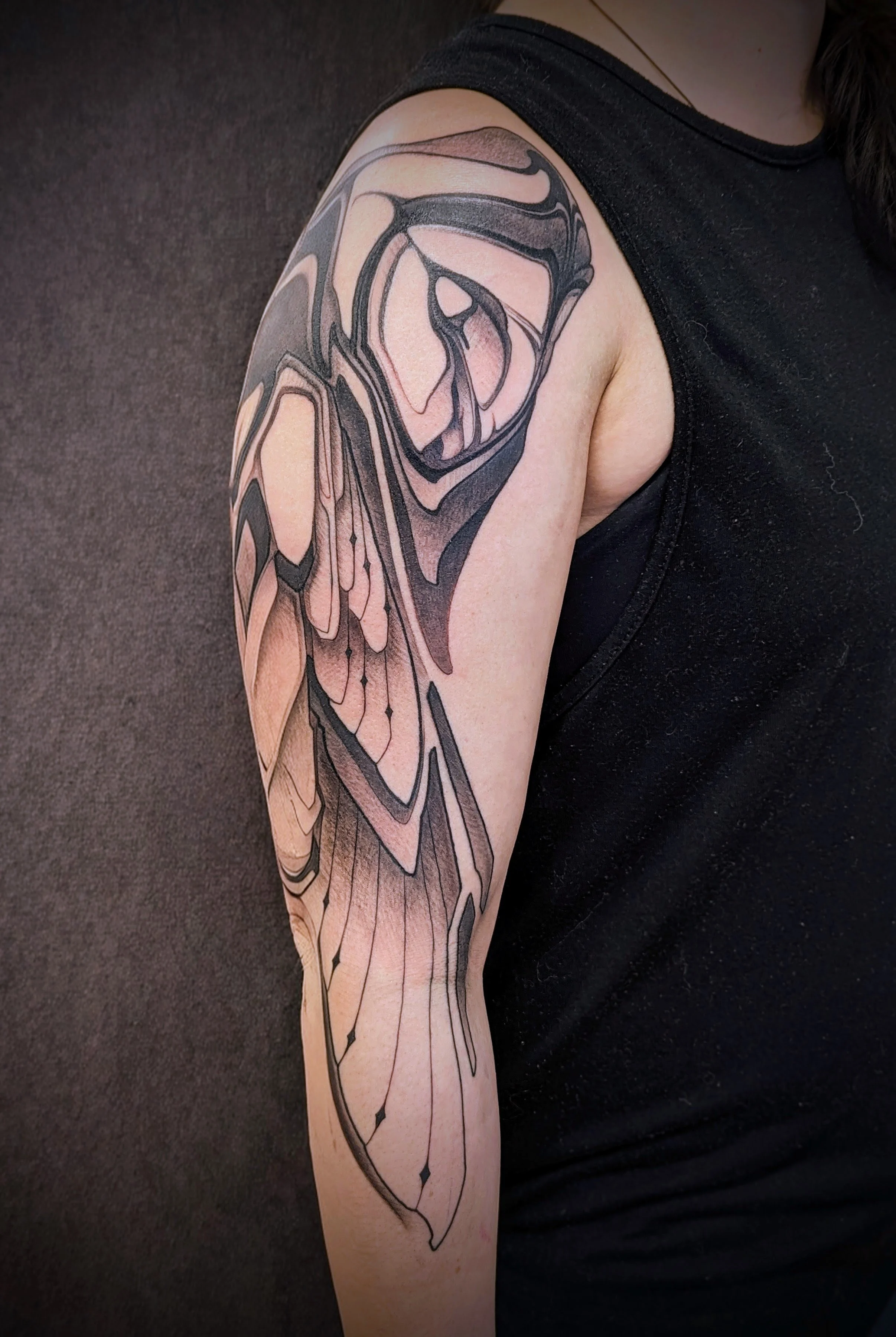 A person's upper arm with a black and peachlineart butterfly tattoo, with detailed wing patterns, against a gray background.