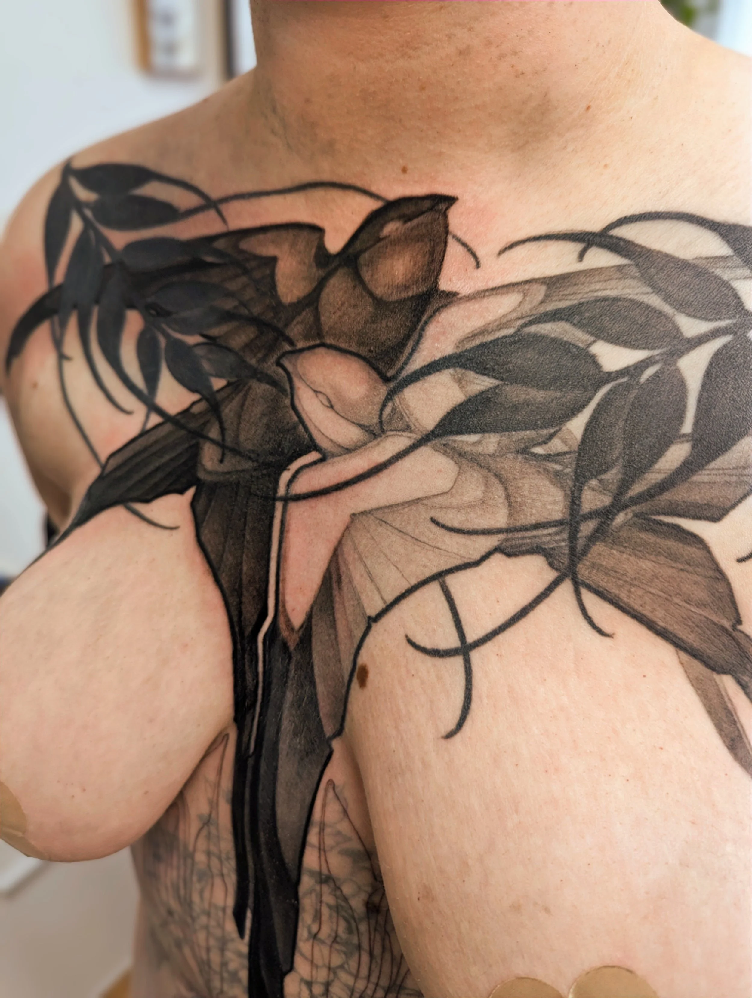 Close-up of a person's shoulder and chest tattoo of a hummingbird with wings spread, surrounded by abstract leaf and branch designs.