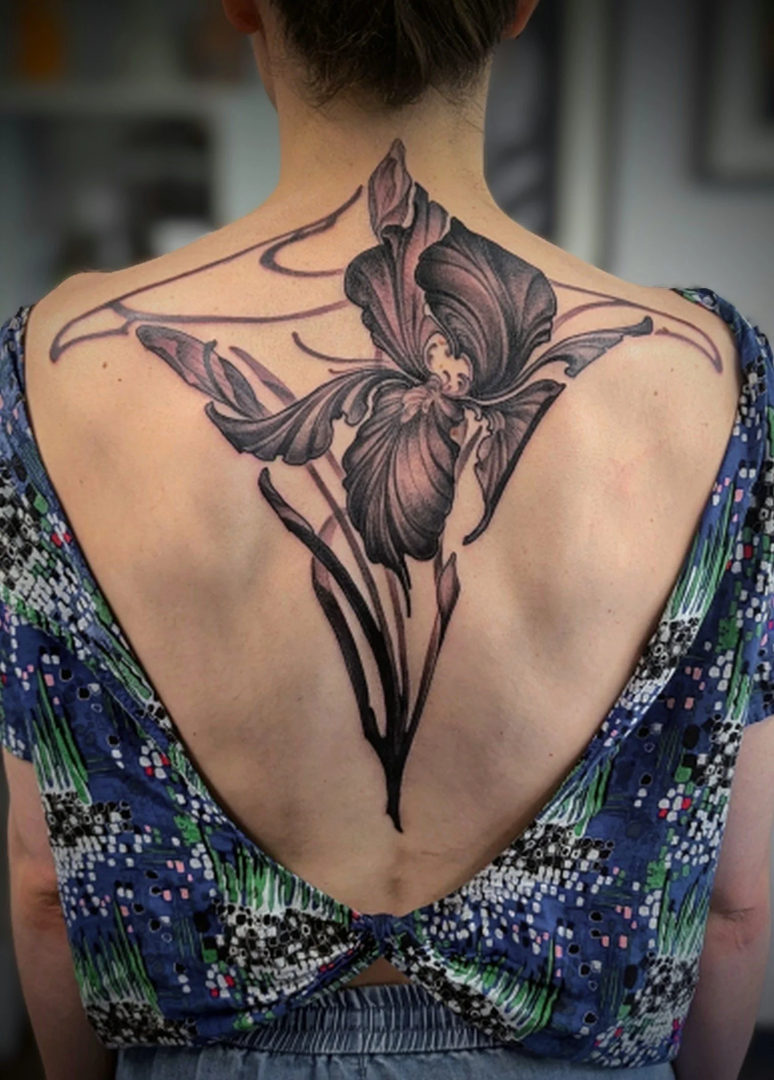 Black and gray tattoo of a butterfly with elongated, floral-like wings on a person's upper back, extending from shoulder to shoulder.