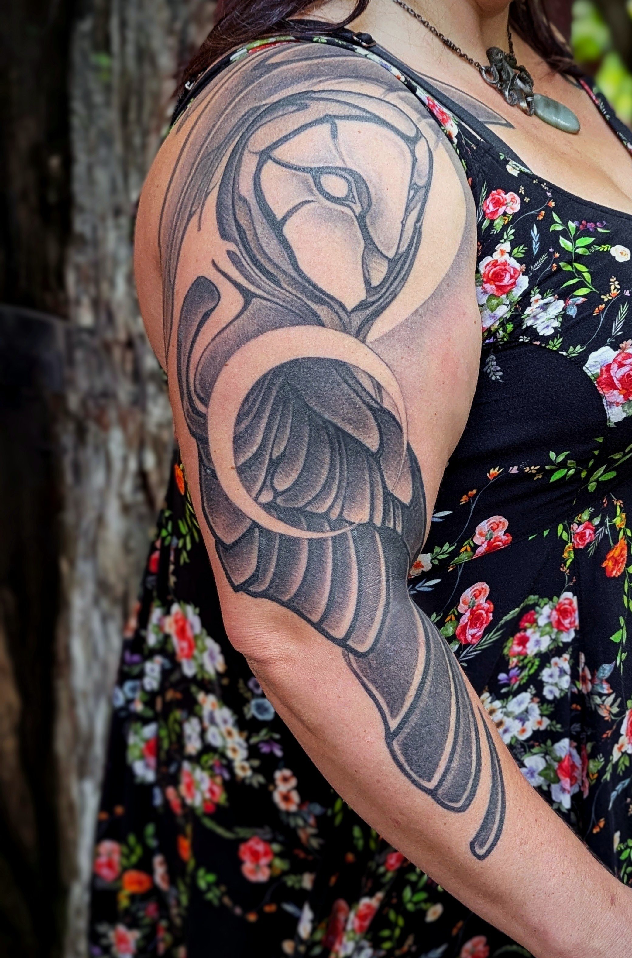 A person with a large, detailed black and gray tattoo of a snake on their upper arm, wearing a black floral dress with red, pink, and white flowers.