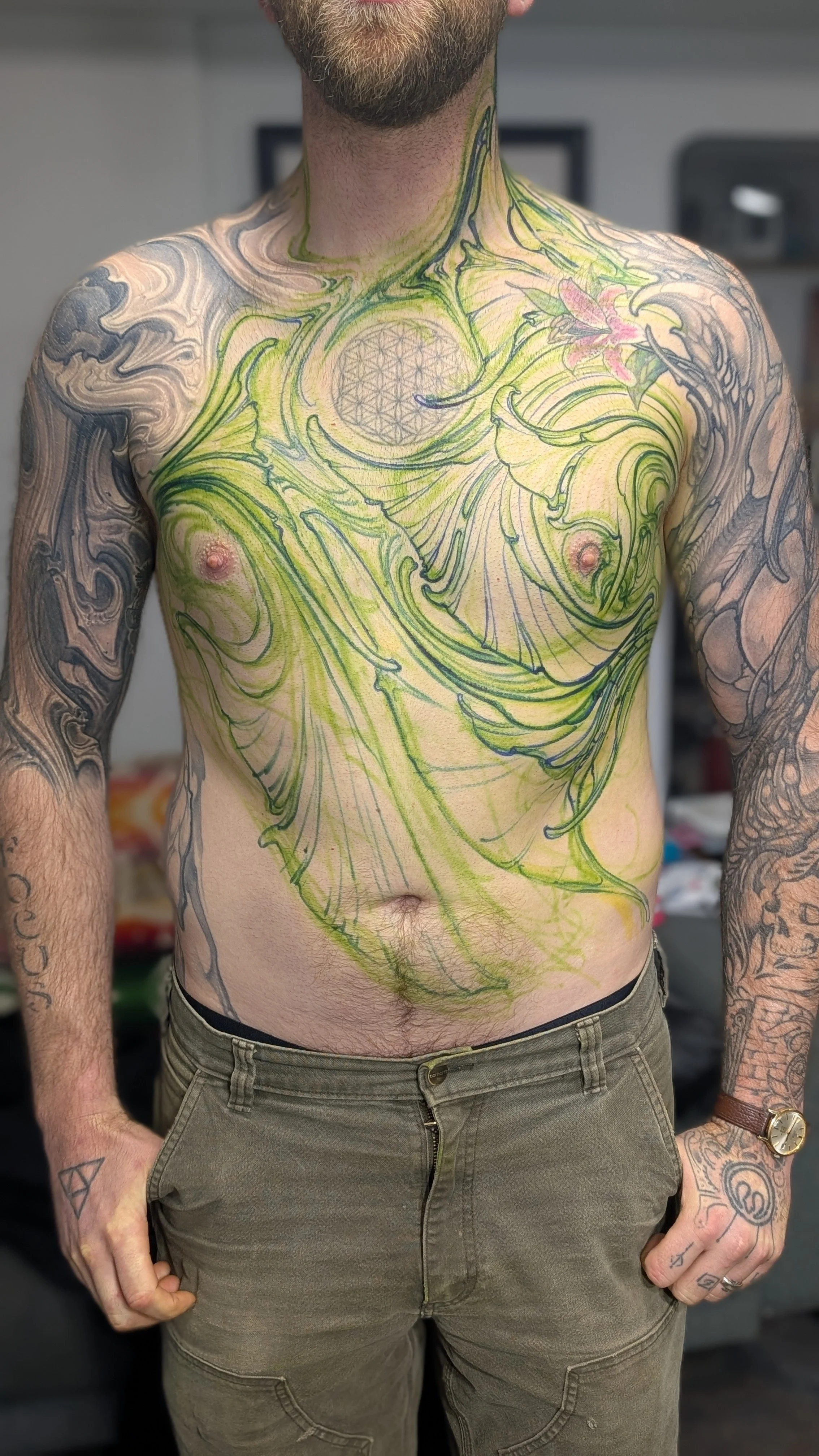 A man with tattoos on both arms and a tattooed chest with green outlines, pink flowers, and geometric shapes. He is wearing brown pants and a wristwatch.