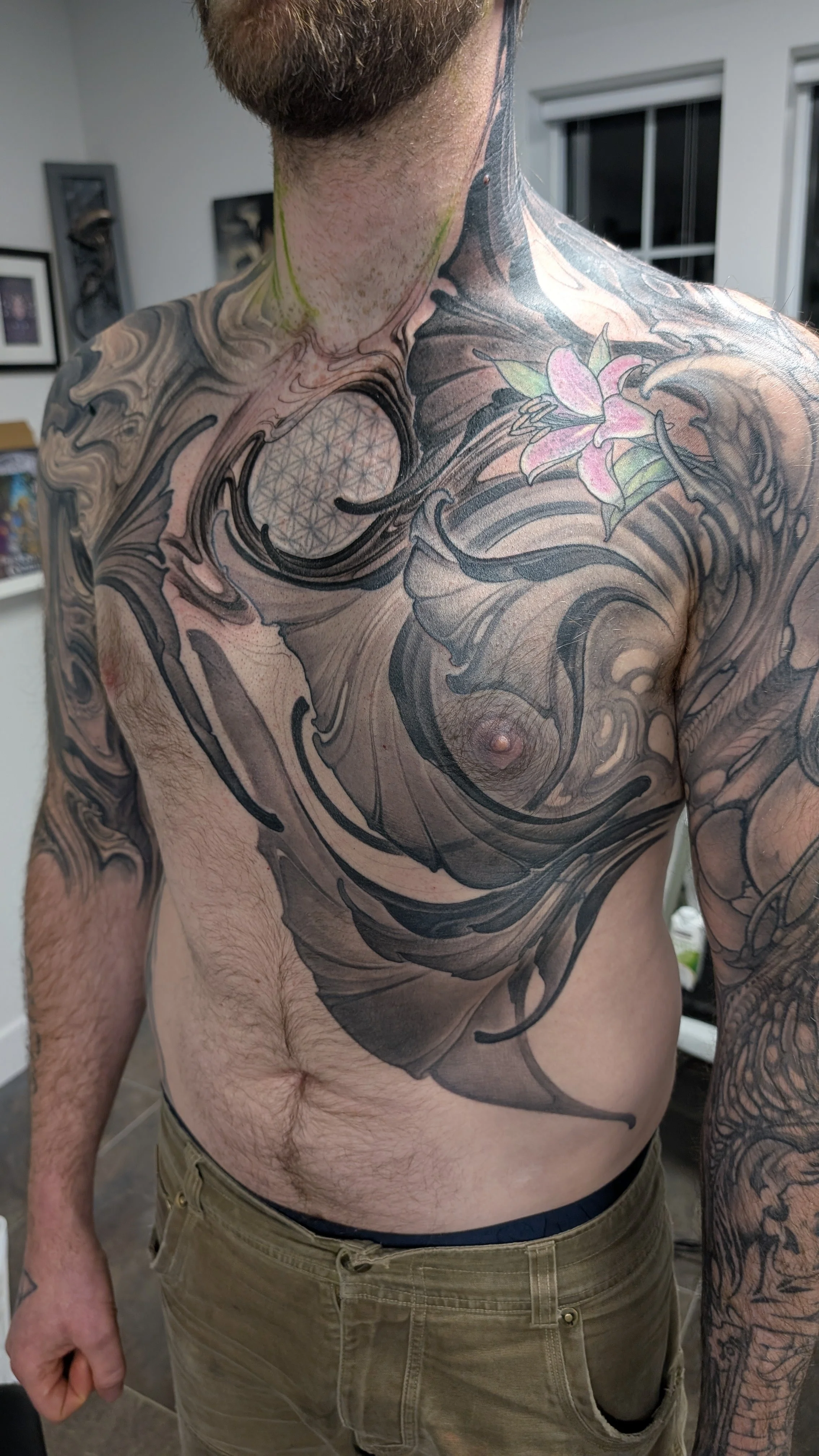 A man with extensive tattooing on his chest, shoulders, and arms. The tattoos include a lotus flower on his right shoulder, a geometric pattern in a circle on his upper chest, and swirling abstract designs covering much of his torso and arms.
