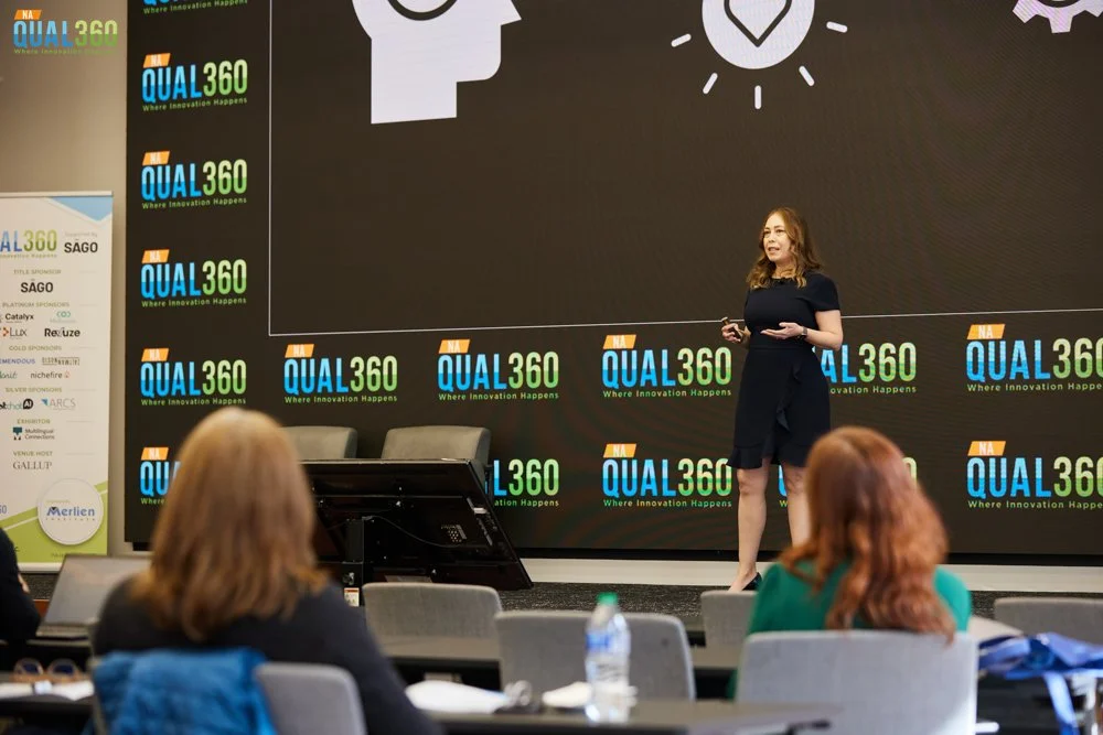 A woman giving a presentation on stage at the Qual360 conference, with a large screen behind her displaying the event logo and icons representing innovation and ideas. Audience members are seated and listening.