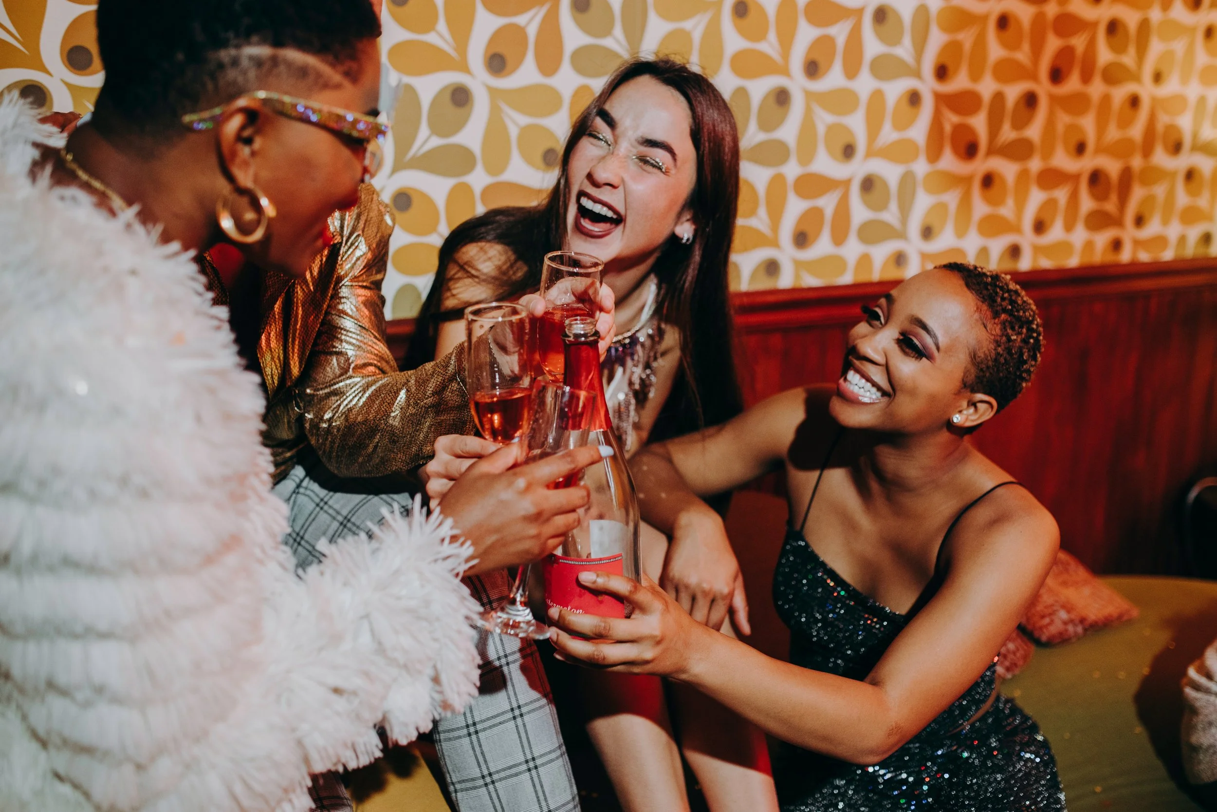 Three women at a party laughing and celebrating with glasses of rosé wine and a wine bottle, in a room with retro patterned wallpaper.