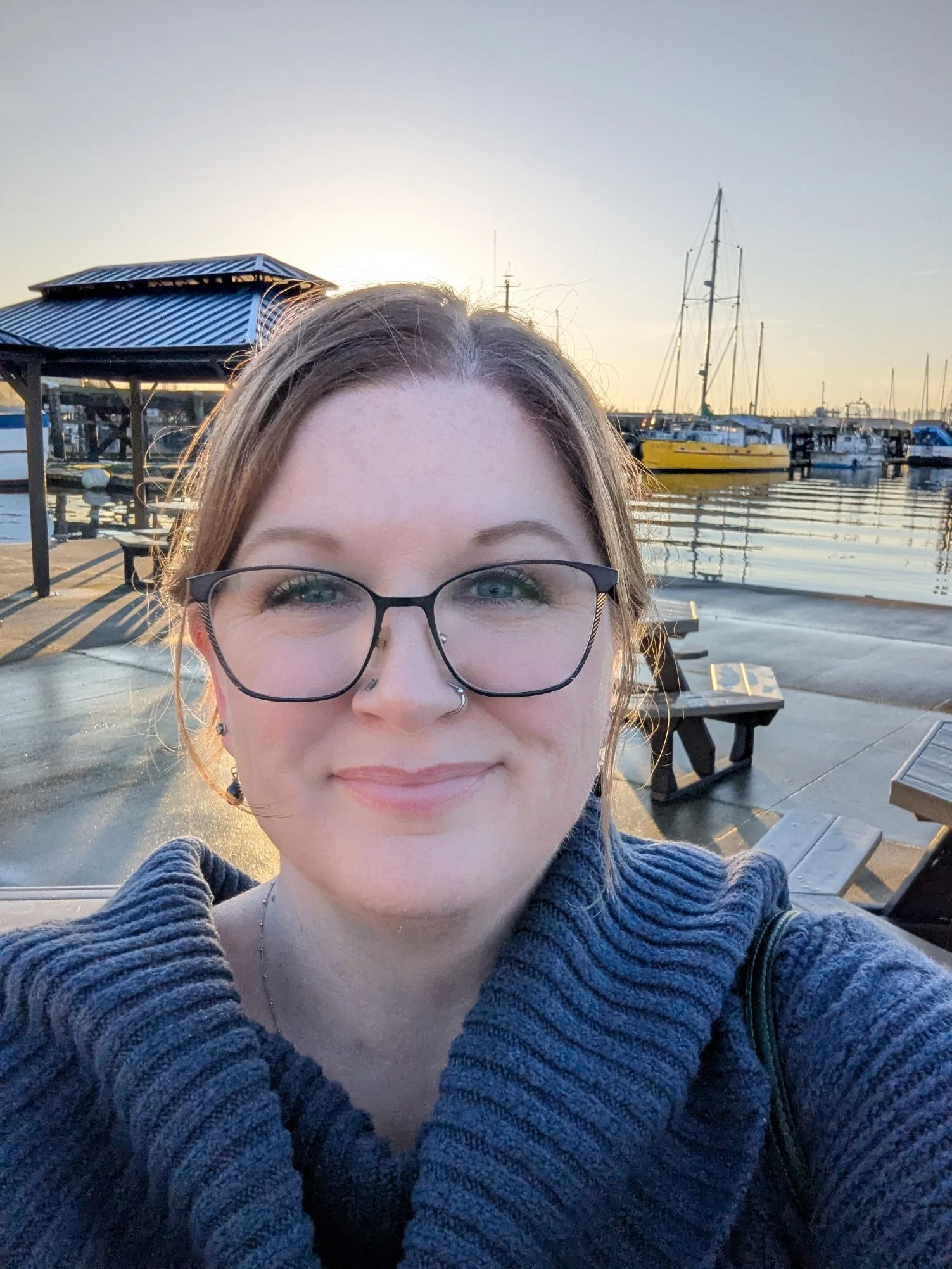 A woman with glasses and a nose piercing taking a selfie at a marina during sunset, with boats and a small pavilion visible in the background.