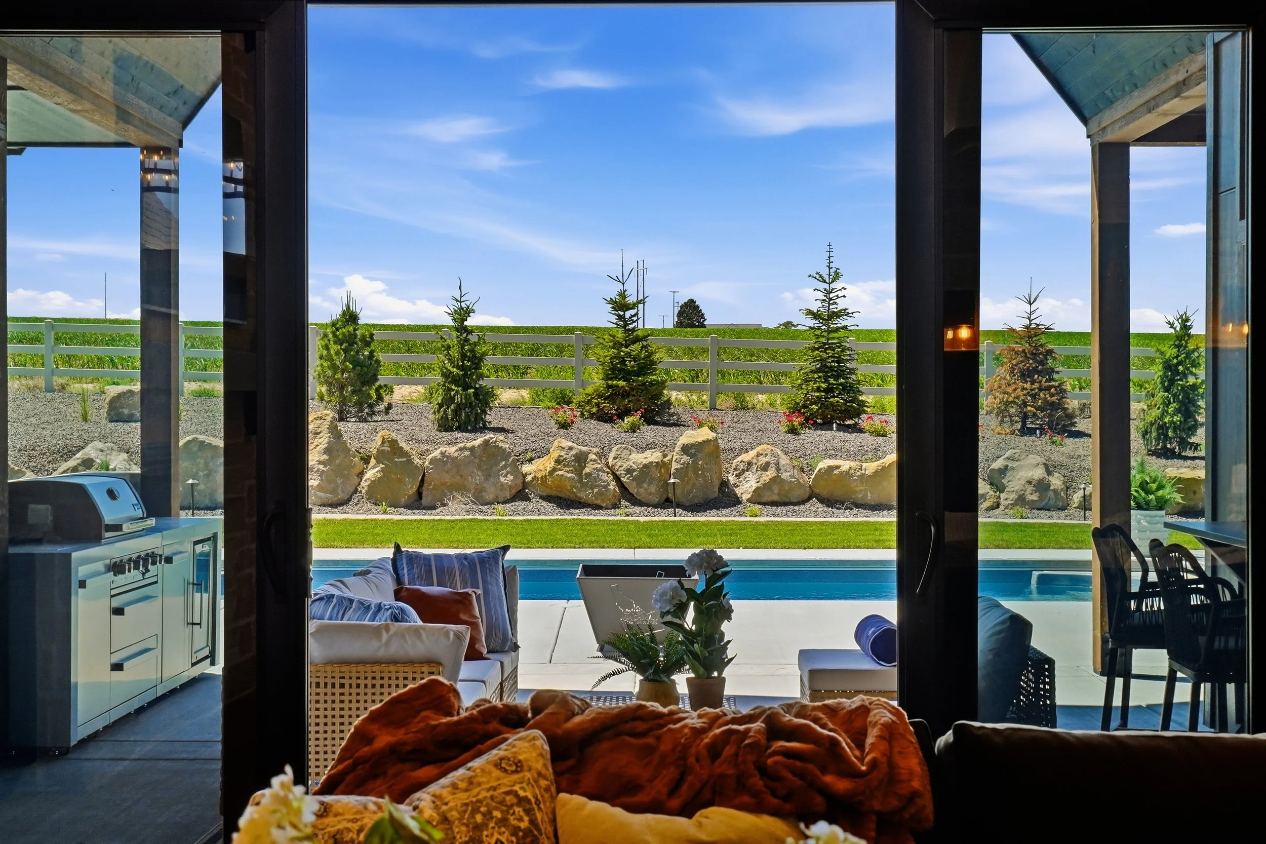 View through open glass doors of The Cody by Bachman Custom Homes, showing a covered patio with outdoor kitchen, lounge seating, and a pool framed by boulder landscaping and countryside views in Lone Star Ranch, Nampa, Idaho.
