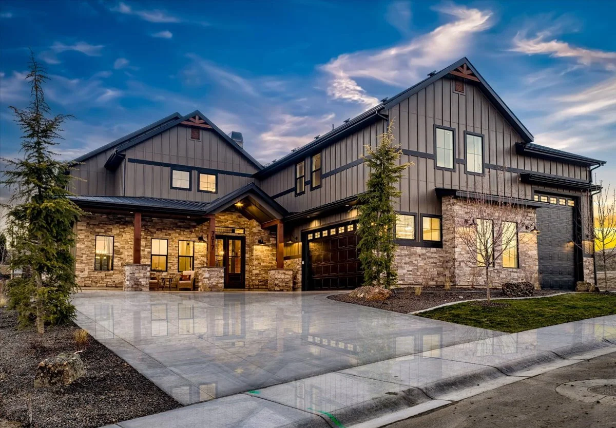 Front exterior view of the Bowers in Valnova. Modern two-story house with stone and dark wood exterior, surrounded by newly planted trees and a smooth concrete driveway, during sunset with cloudy sky.