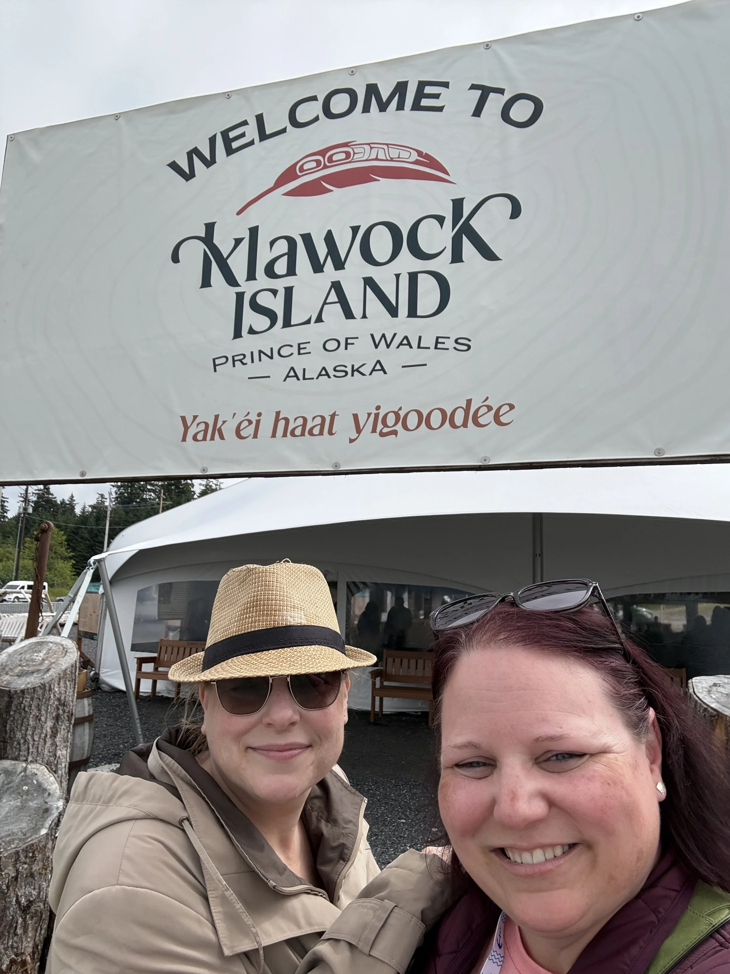 Two women standing in front of the Welcome to Klawock sign