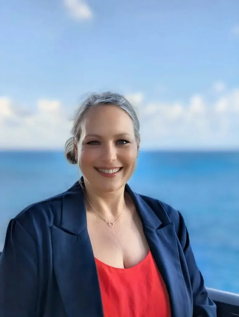 Wendy Pochop, co-founder of Untethered Voyages, standing in front of a bright blue ocean and sky in a red shirt and navy blazer.
