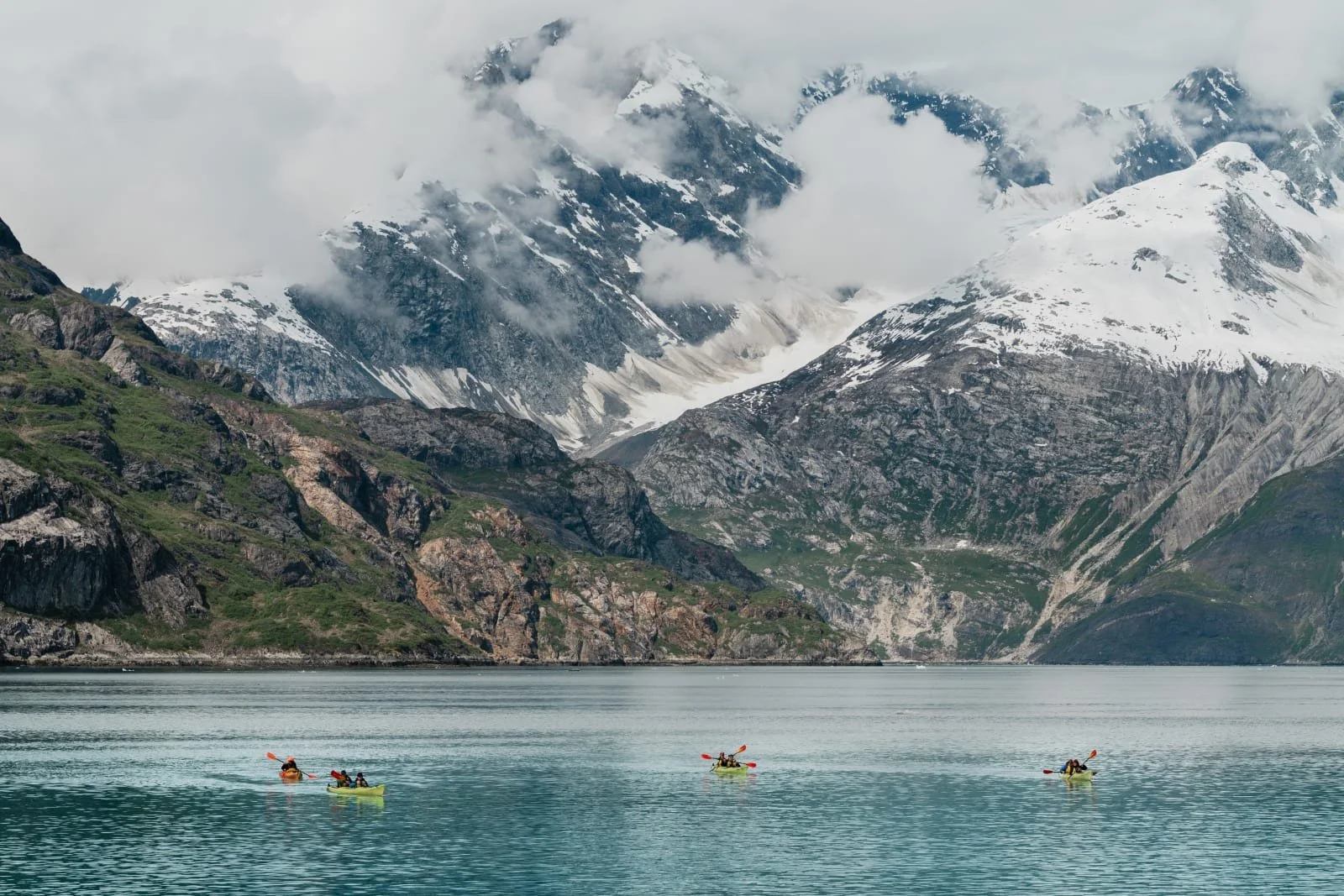 Sea kayakers paddling toward a glacier face in Southeast Alaska, the ice wall towering above them in a remote fjord