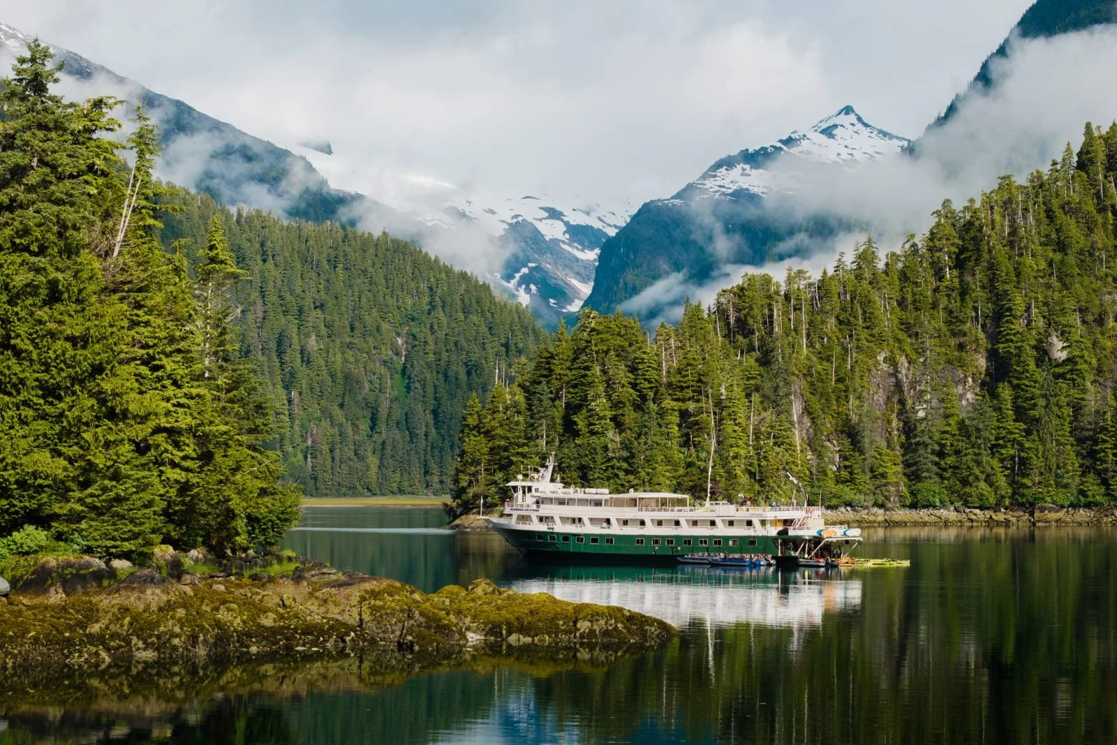 Small expedition ship anchored in a remote Southeast Alaska fjord, surrounded by forested mountains and calm water, with a Zodiac tender in the foreground