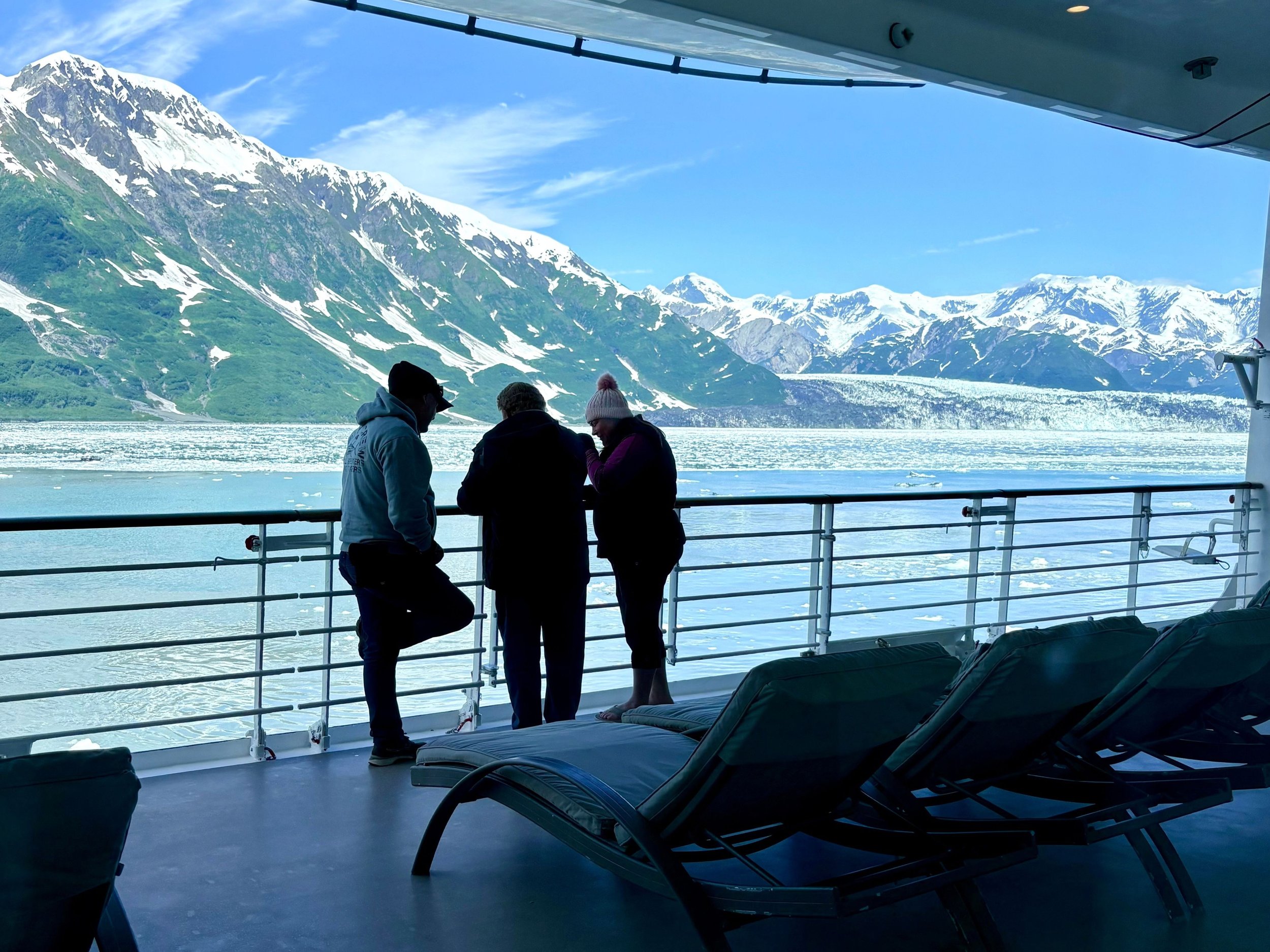 Three passengers look out from a cruise ship deck toward the Alaska coastline