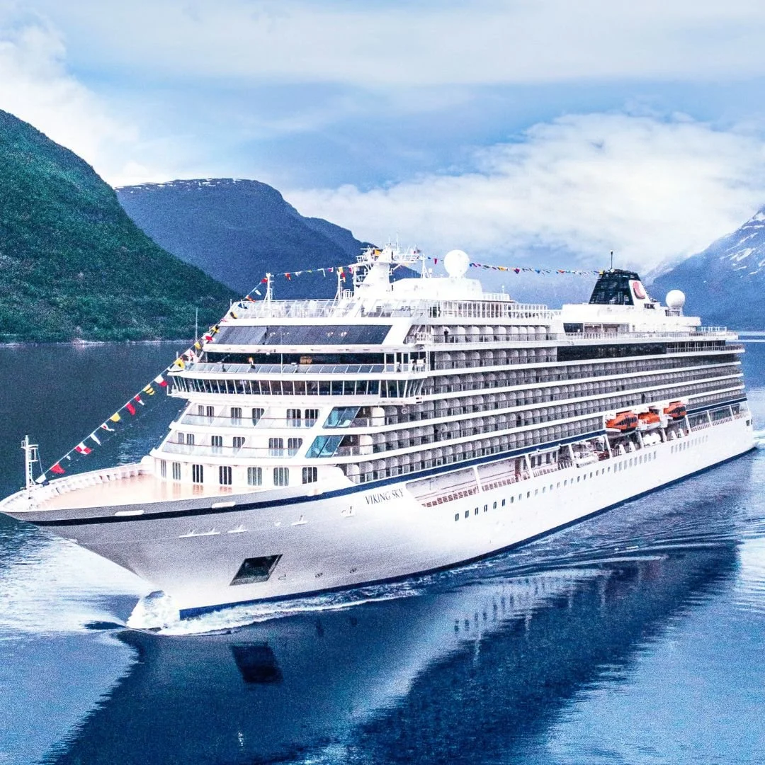 A large white cruise ship sailing through fjord waters with mountains in the background.