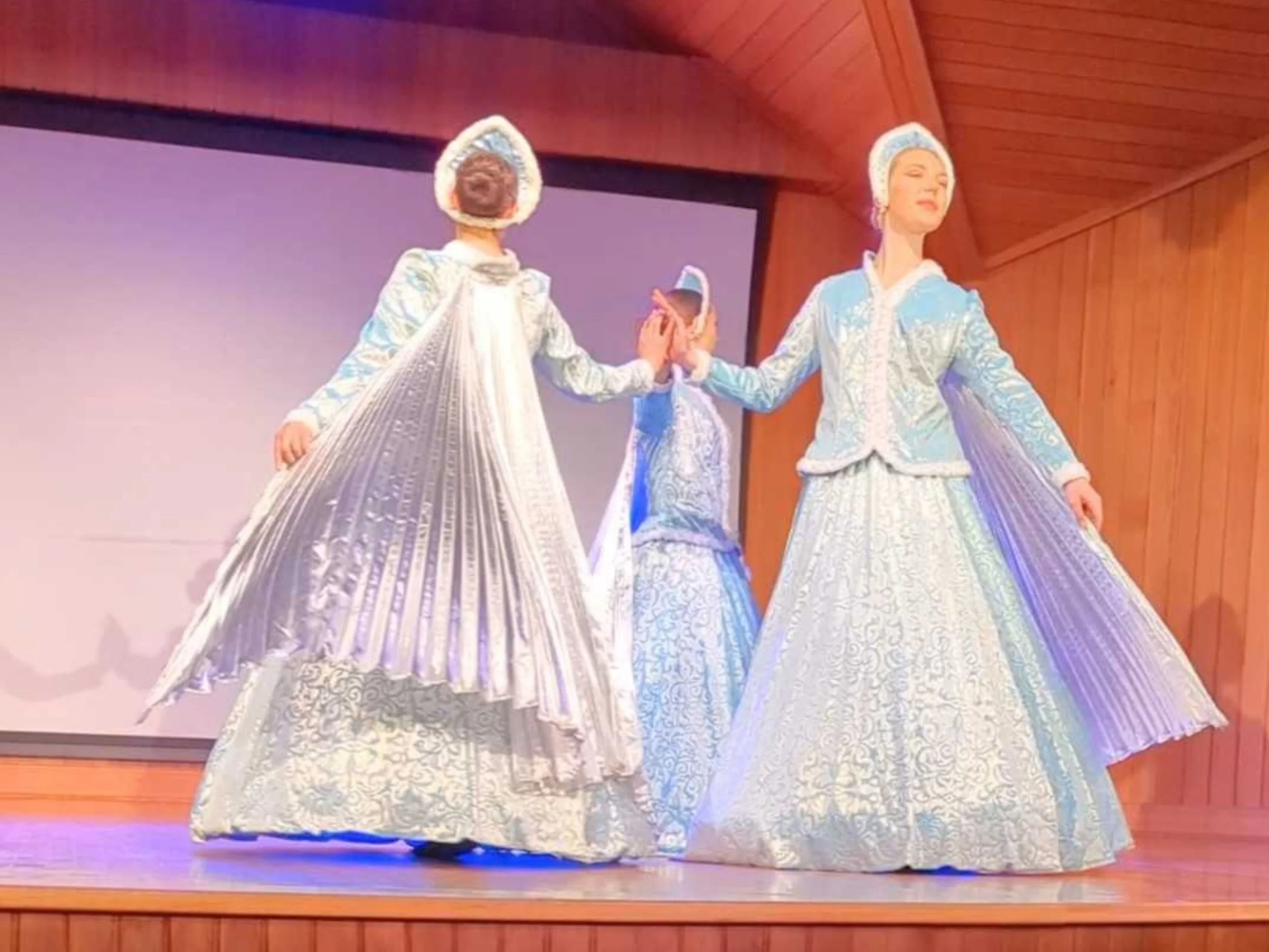 Three women dressed in authentic Russian 1800s dresses and headwear, dancing on a stage.