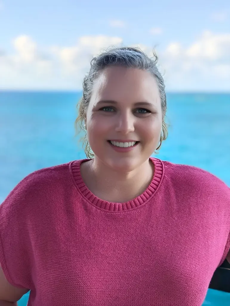 Elizabeth Pochop, co-founder of Untethered Voyages, standing in front of a bright blue ocean and sky in a vibrant hot pink sweater.