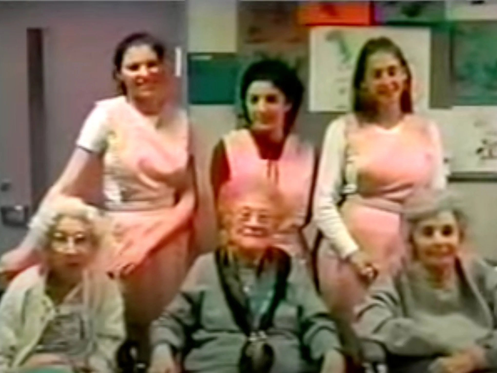 Six people, three standing women in pink and white outfits, and three seated children wearing glasses or caps, in an indoor setting with posters on the wall.