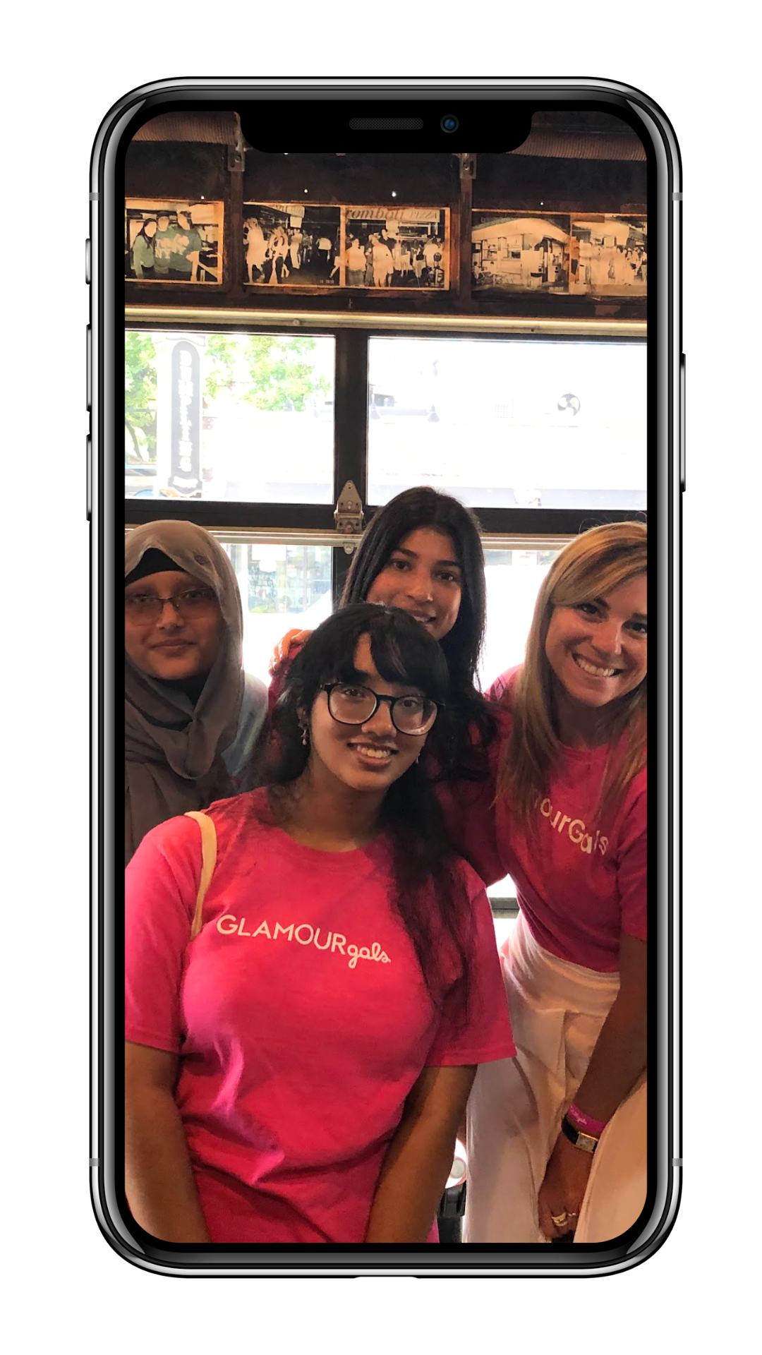 Four women smiling inside a restaurant, two wearing matching pink GLAMOURGALS t-shirts, posing for a group photo.