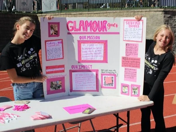 Two young girls standing on a track field holding a pink and white project board about Glamour Girls. The board has sections titled 'Our Mission', 'Our Impact', and 'Service Hours', with pink and white paper details. The girl on the left wears a black t-shirt with white design, and the girl on the right wears a black shirt. There are pink paper items on the table in front of them.
