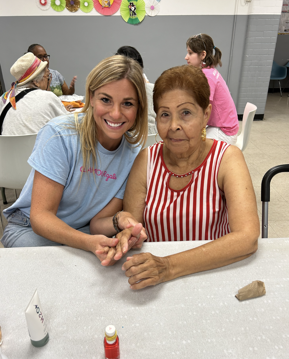 Two women, one young with blonde hair and an older woman with red hair, sit together at a table holding hands and smiling. Behind them, there are other women seated at the table and colorful paper decorations on the wall.
