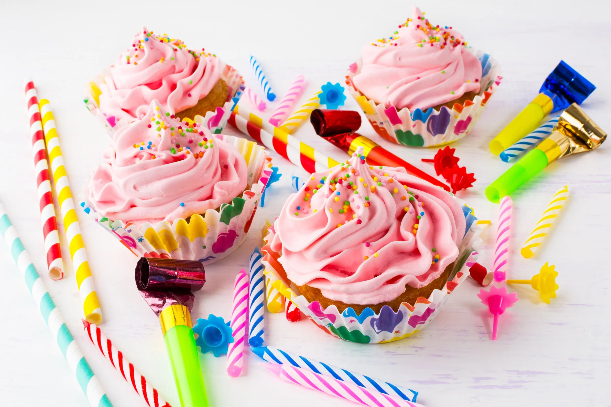 Pink frosted cupcakes with colorful sprinkles surrounded by striped birthday candles and party noisemakers on a white surface.