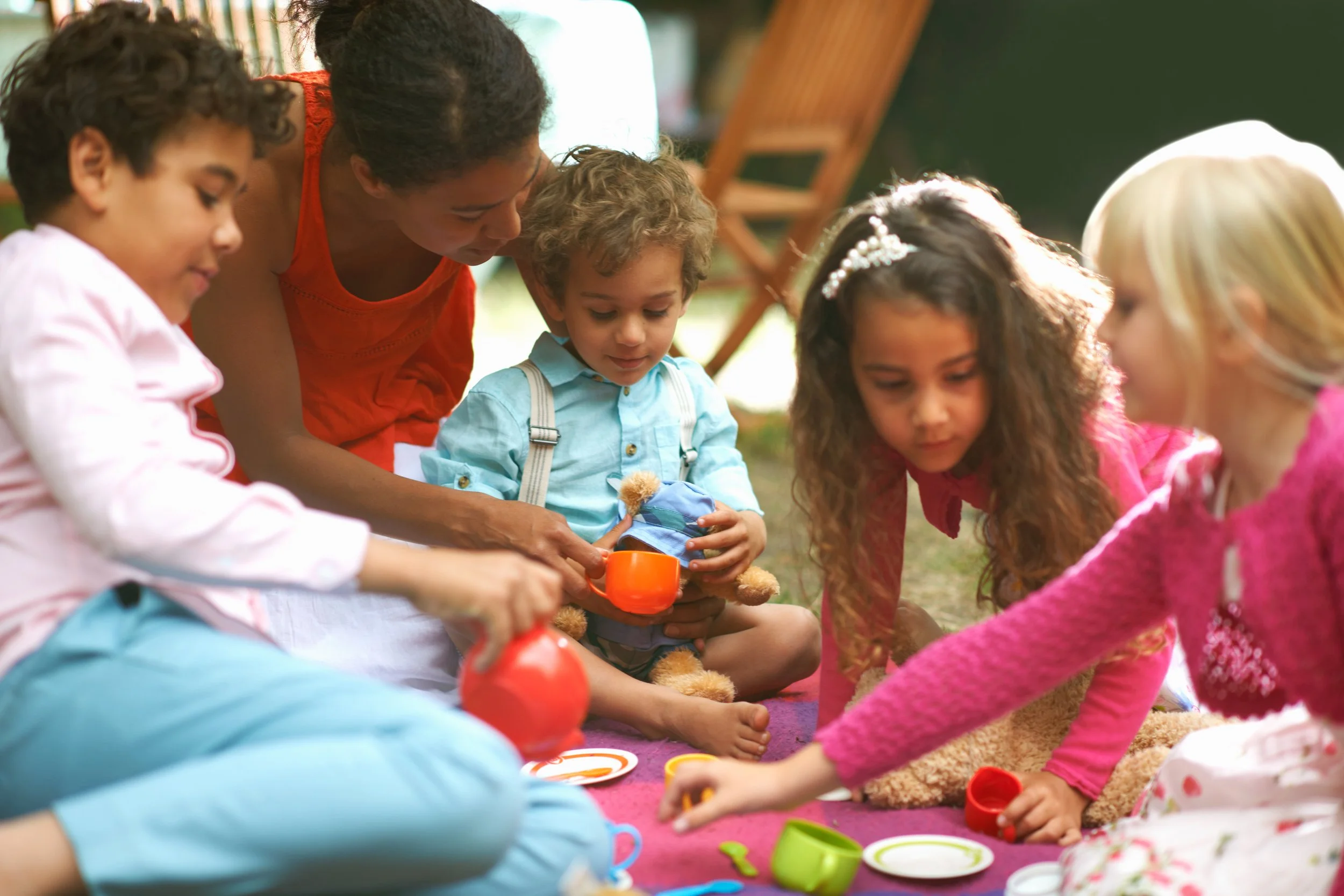A group of children and an adult having a tea party outdoors, with tea cups, plates, and a teddy bear.