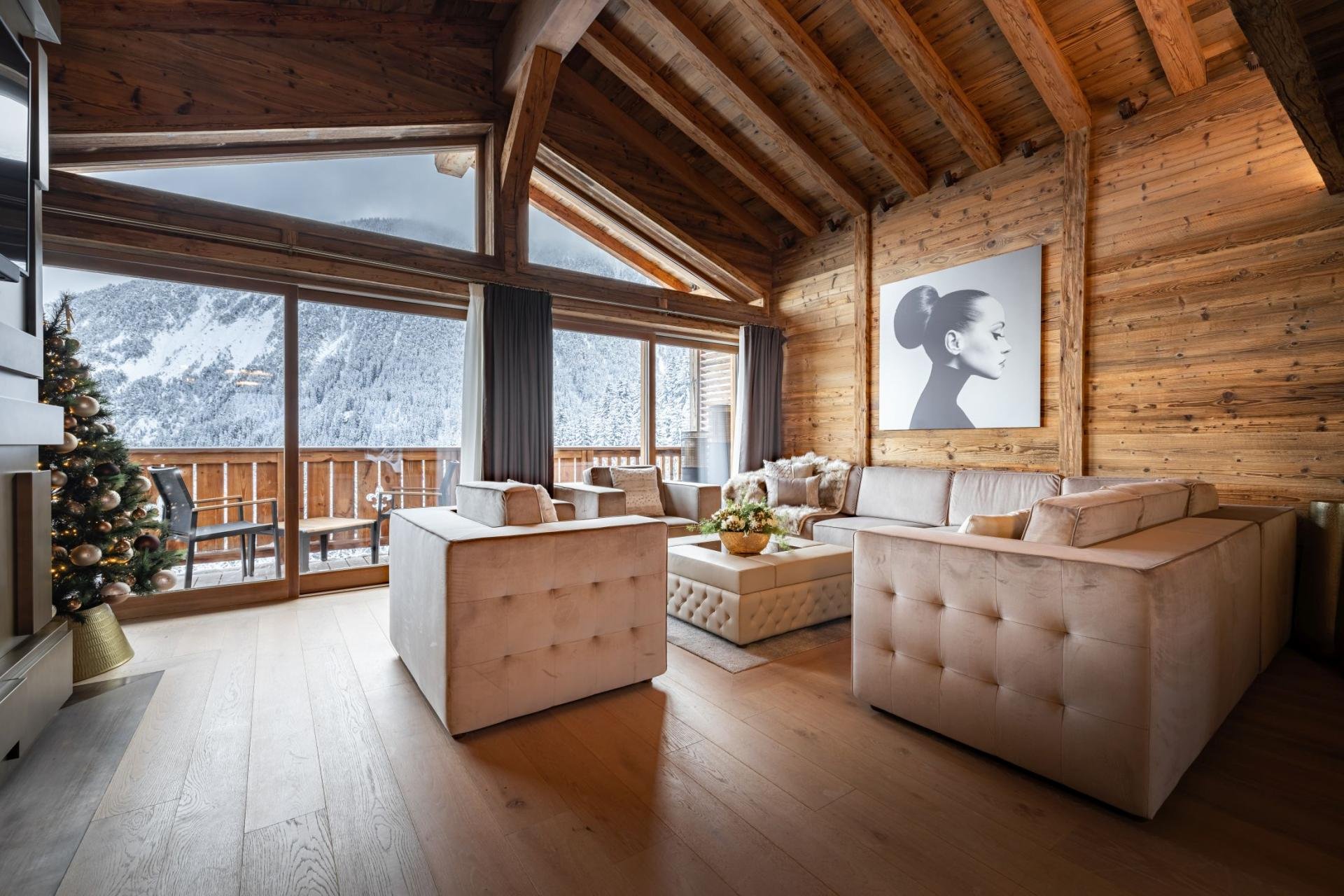 Living room with beige sofas, wooden walls and ceiling, large windows showing snowy mountains, a Christmas tree with ornaments, and black and white portrait wall art.