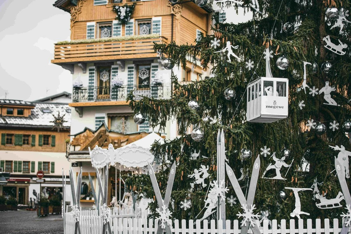 Decorated Christmas tree in Megeve with white and silver ornaments, snowflake and skater cutouts, and a small gondola. In the background are traditional alpine-style buildings.