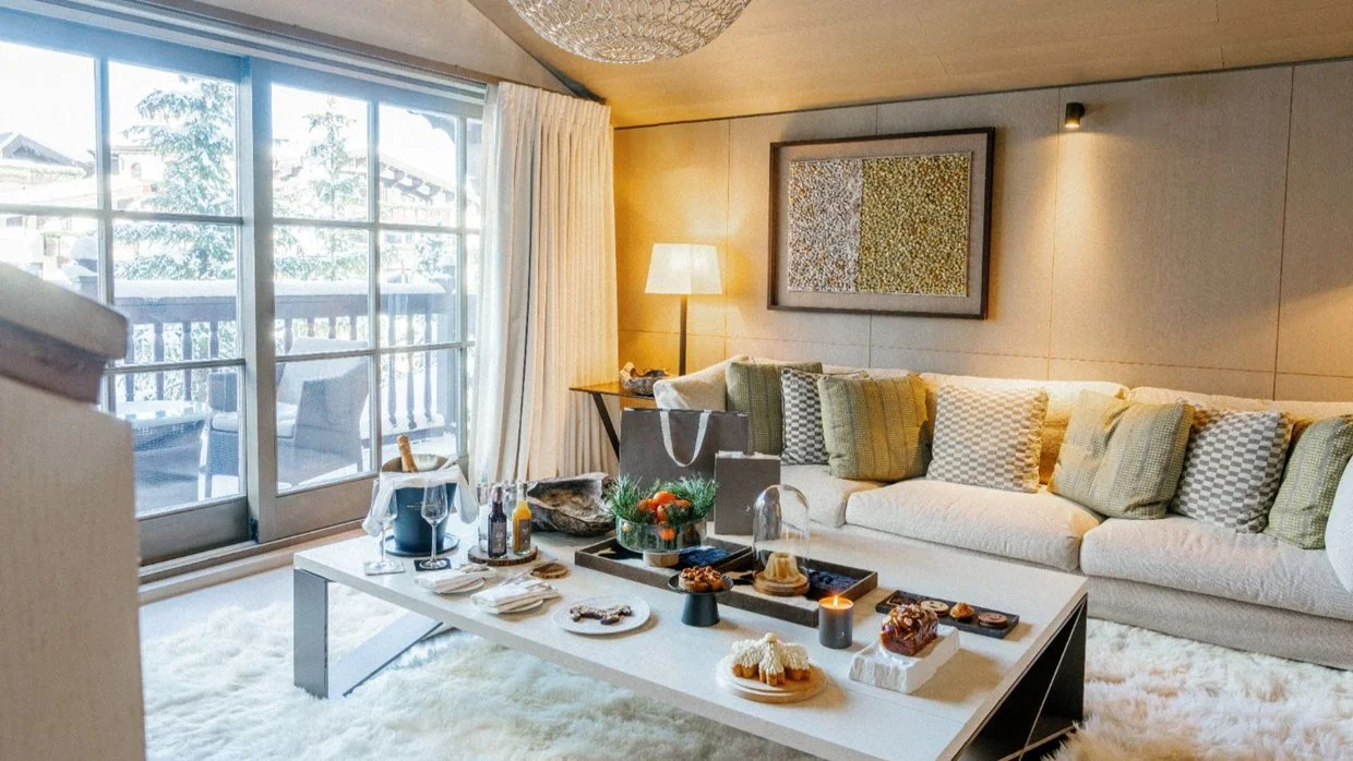 Living room with beige sofa, green and checkered cushions, floor lamp, artwork, coffee table with snacks, drinks, and holiday decorations, sliding glass door leading to balcony with outdoor furniture, and a white fluffy rug.