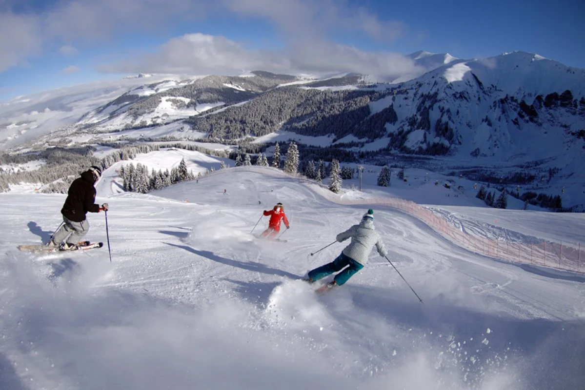 Three skiers skiing down a snowy mountain slope in Megeve with snow-covered trees and mountains in the background on a partly cloudy day.