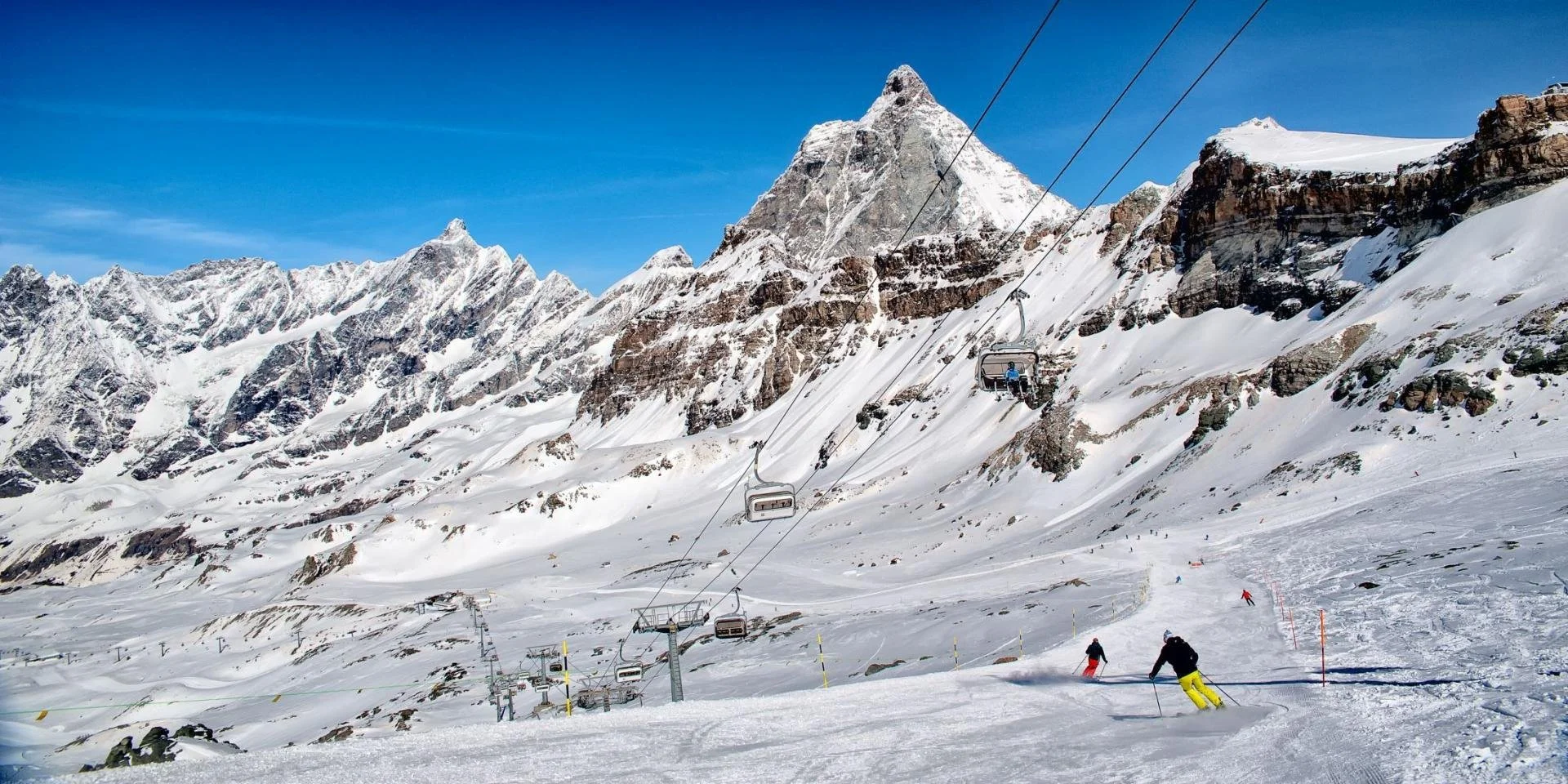 Snow-covered ski slopes with skiers and a ski lift, set against a backdrop of rugged, snow-capped mountains under a clear blue sky.