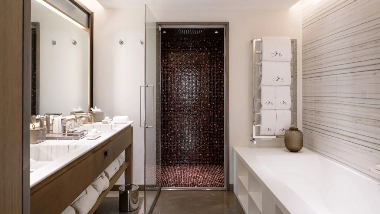Modern hotel bathroom with double sink vanity, large mirror, glass shower with dark mosaic tiles, white towels on a rack, decorative vase on white countertop, and light-colored textured wall paneling.