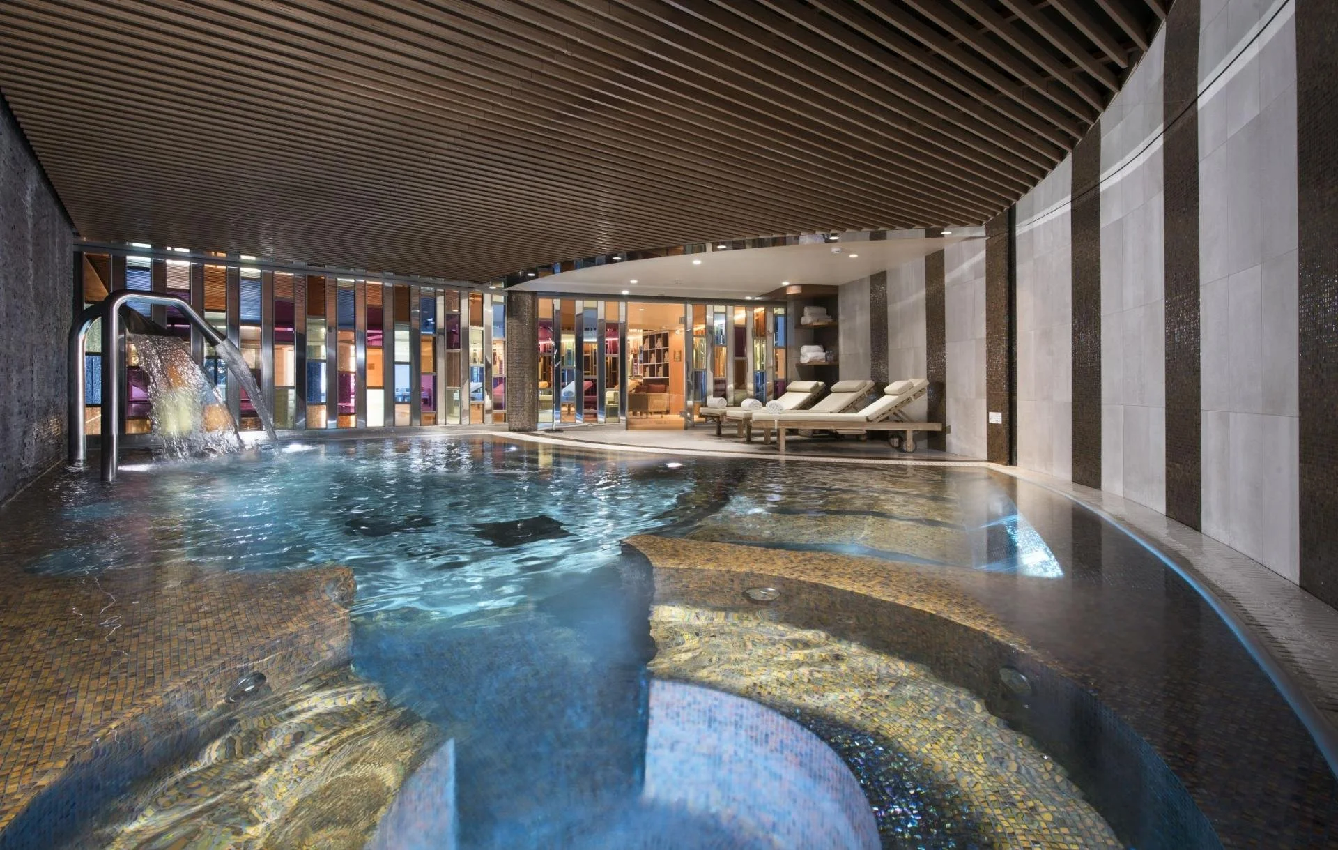 Indoor spa pool area with loungers and towels, wooden ceiling, colorful glass wall, and a water feature.