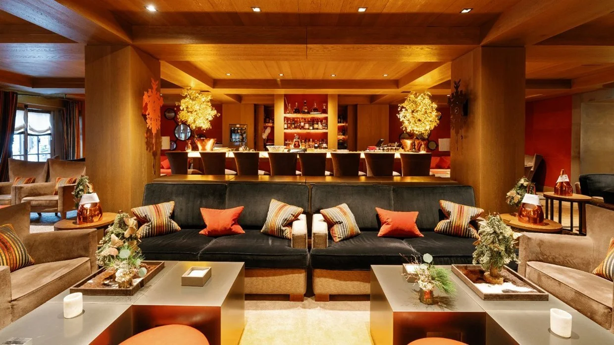 A cozy living room with a large black sofa, colorful striped and red throw pillows, and small tables with decorative items like candles and plants. In the background, there is a bar area with bar stools, warm lighting, and wooden accents, creating a 