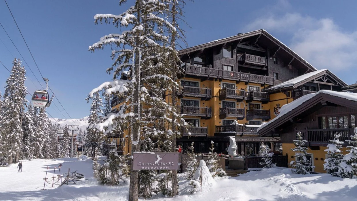 Snow-covered alpine hotel with balconies and a ski lift to the left, surrounded by snow-laden trees, in a winter scene.