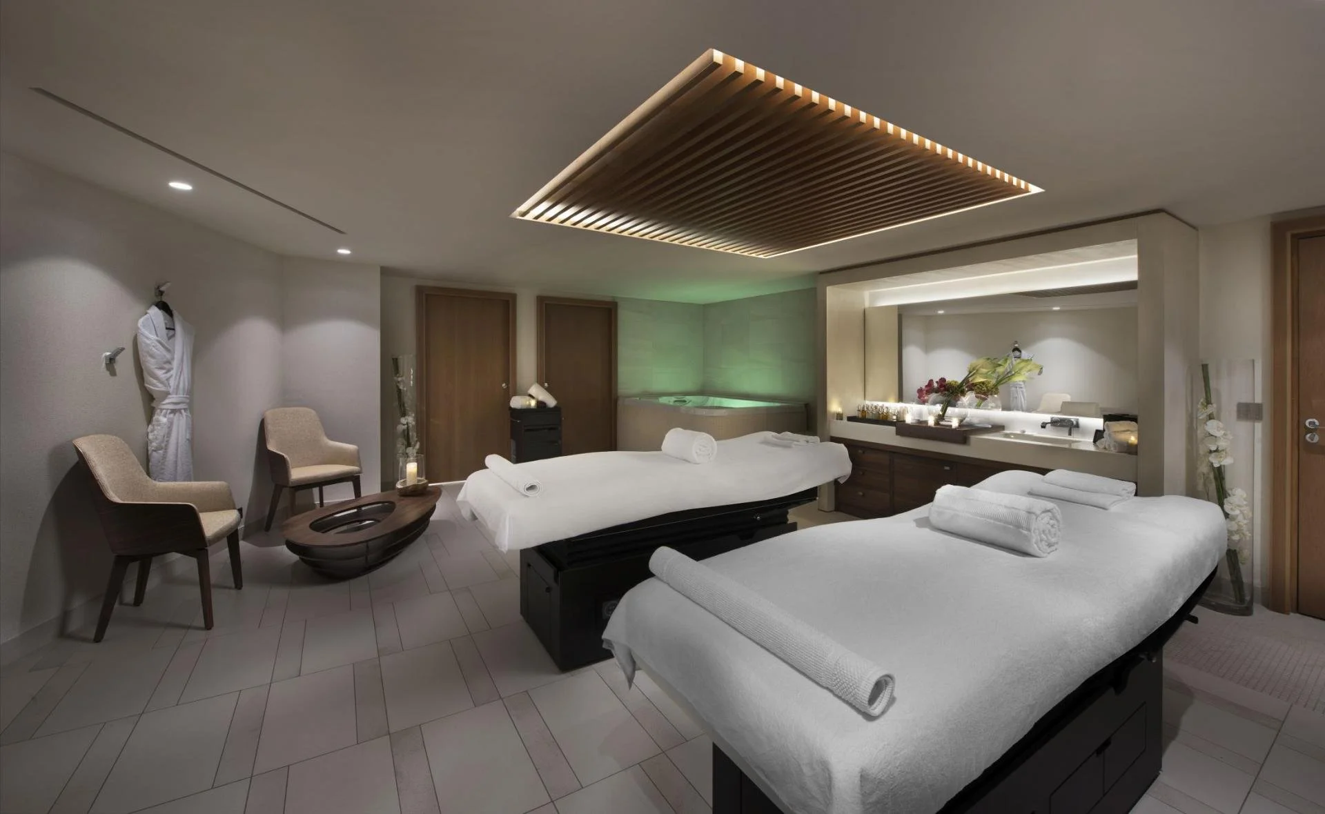 A luxurious spa or massage room with two massage tables draped in white linens, each with a rolled towel on top. The room features modern decor with neutral tones, a wooden ceiling panel, chairs, decorative vases with flowers, and a small hot tub in 